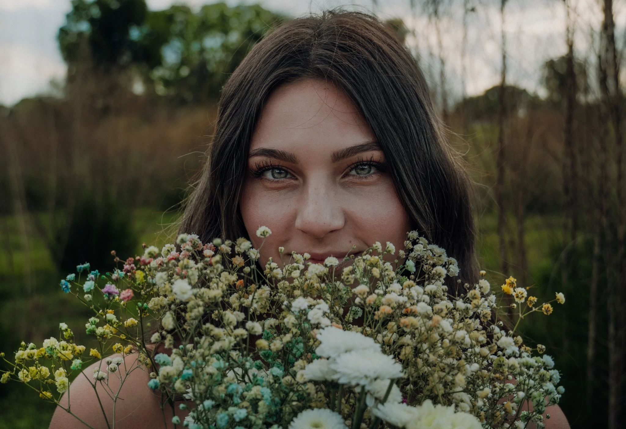 A woman with long dark hair and blue eyes holding a large bouquet of multicolored small flowers outdoors.