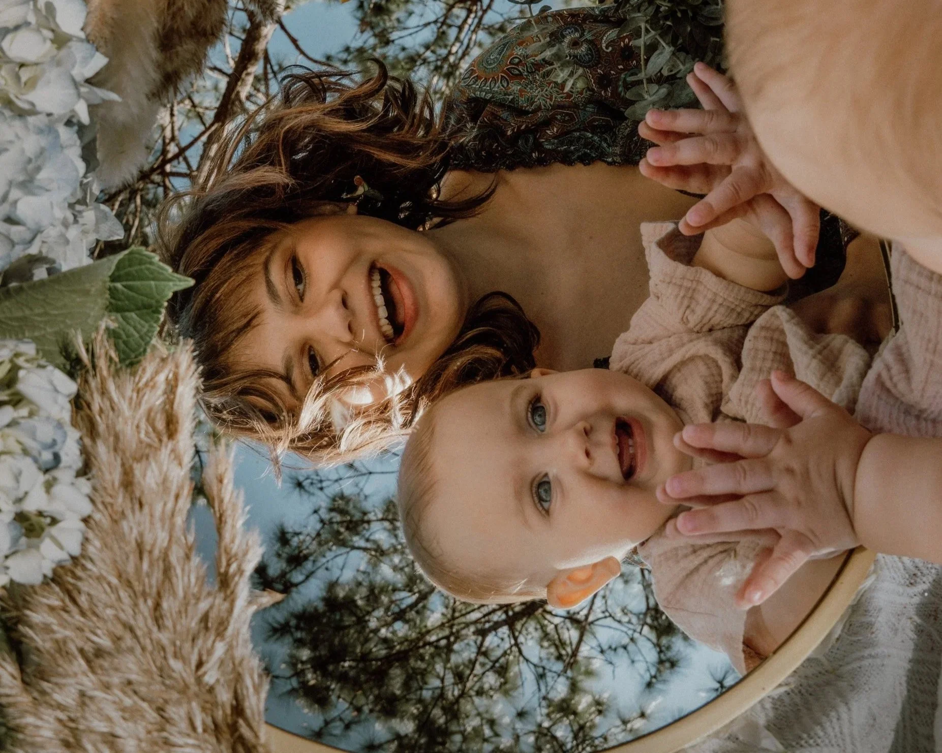 A woman with wavy brown hair and a young girl with blonde hair are smiling and making peace signs with their hands while looking into a round mirror. The scene appears to be outdoors, with trees and a cloudy sky in the background. There are decorative dried plants and white flowers at the edge of the mirror.