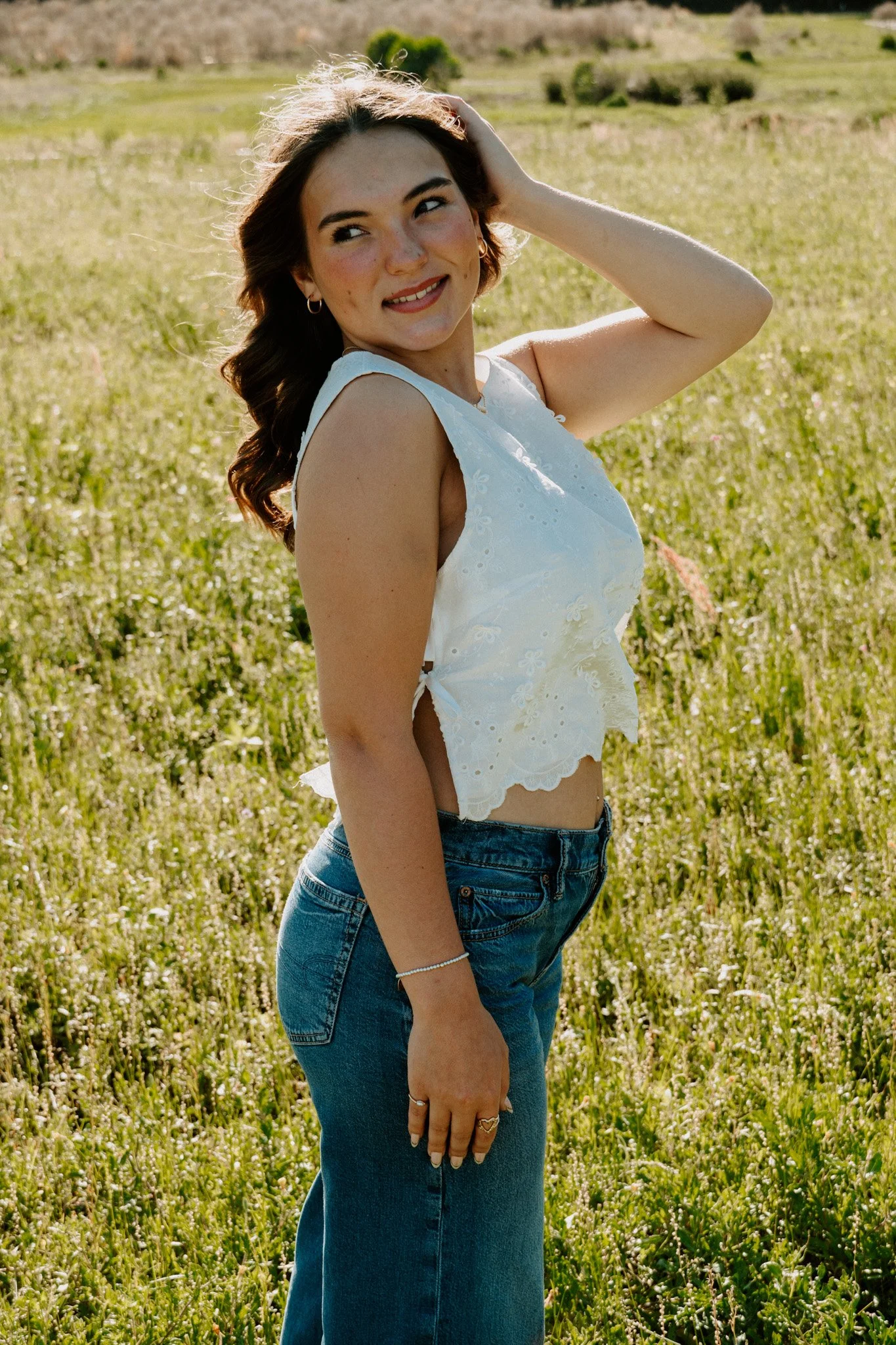 A young woman with brown hair, wearing a white sleeveless top and blue jeans, standing in a grassy field with hills in the background, smiling and looking to her left with sunlight illuminating her face.