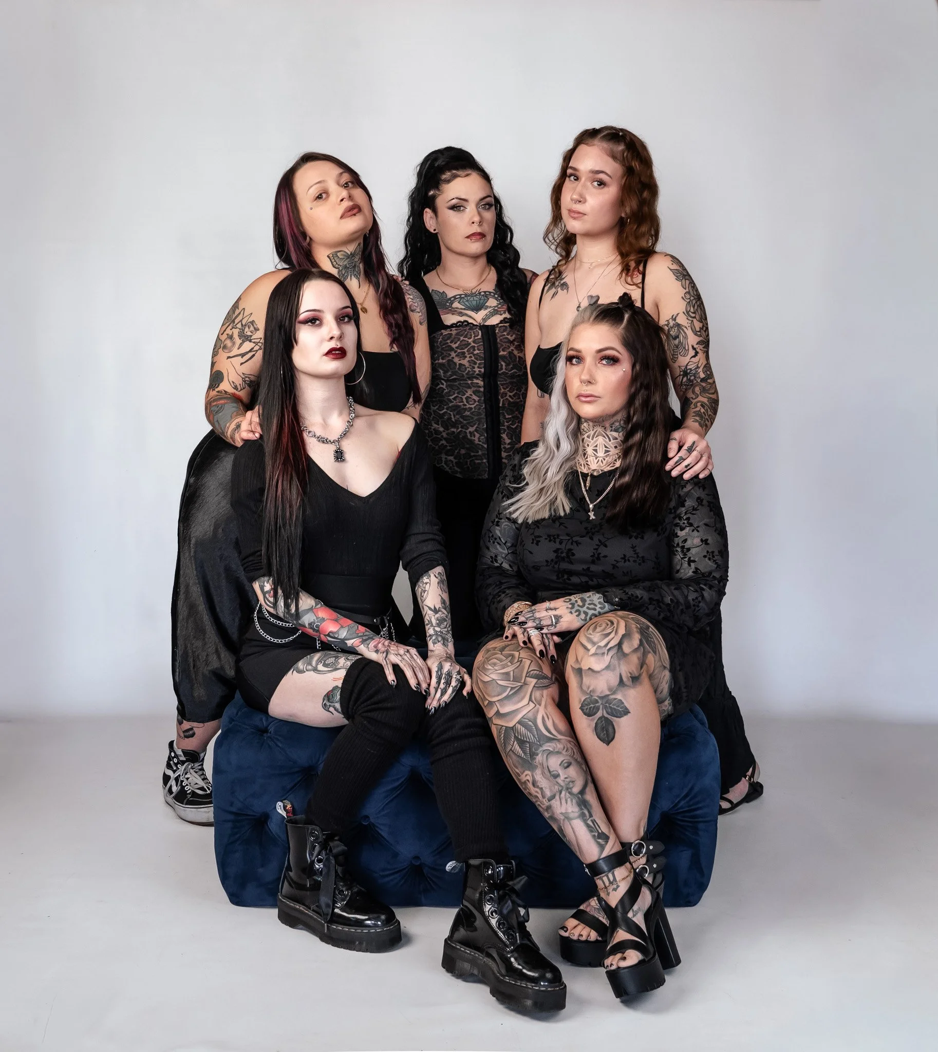 Group of six women with tattoos in black clothing posing together in a studio. Two women are seated, and four are standing behind them. They have various hairstyles and makeup.