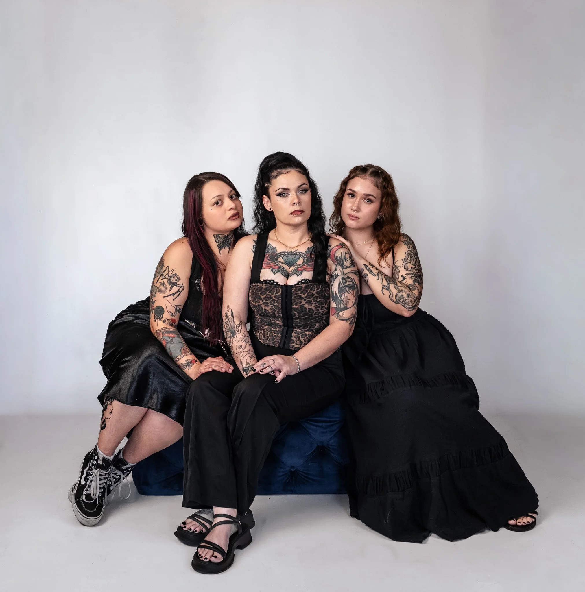 Three women with tattoos sitting on a dark blue couch against a plain white background, dressed in black clothing, with a confident and serious demeanor.