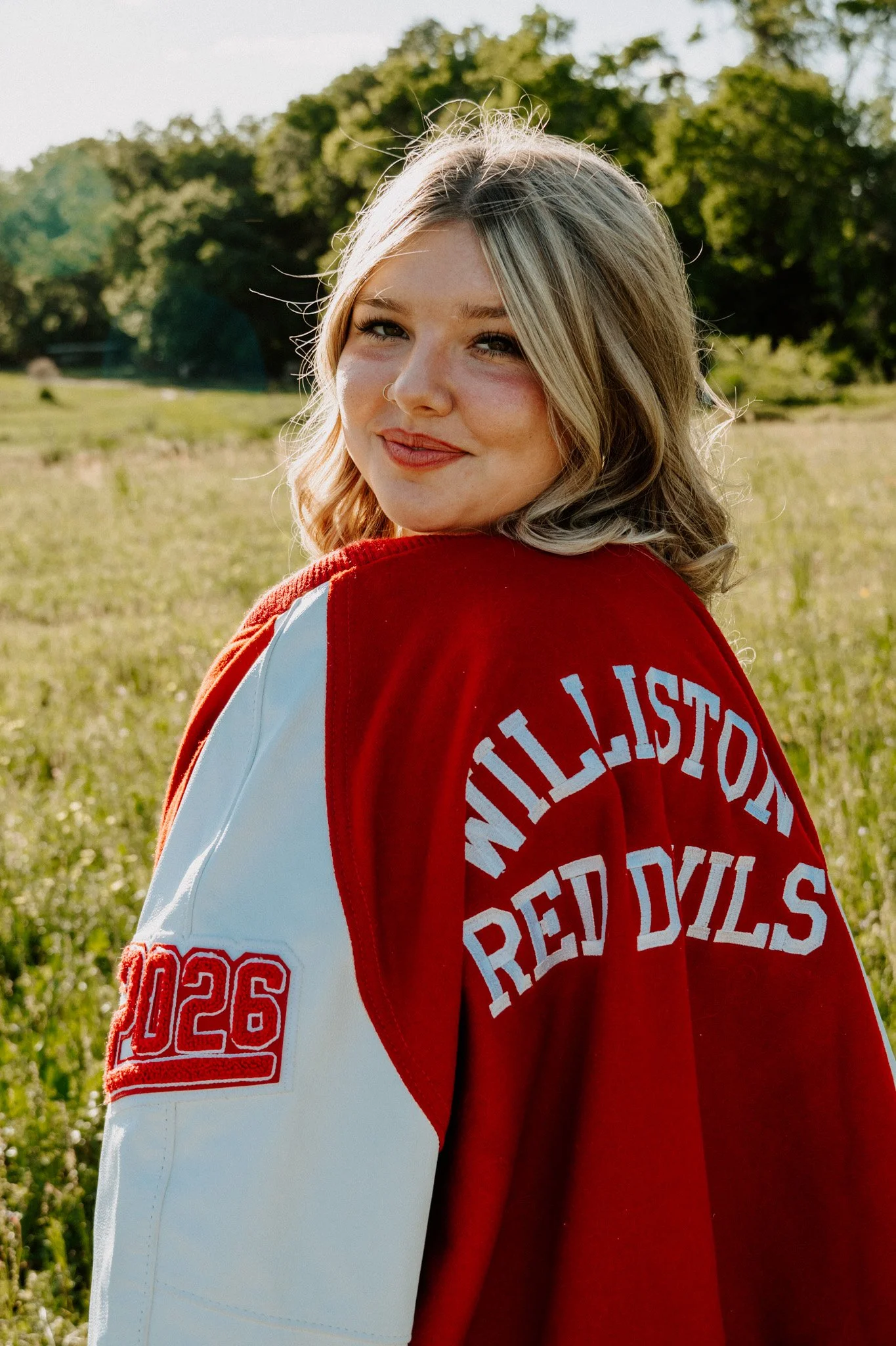 A young woman with shoulder-length blonde hair, wearing a red and white varsity jacket, smiling at the camera outdoors during daylight. The background features green grass and trees.