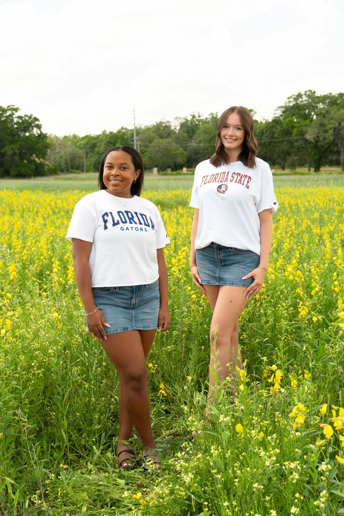 Two young women standing in a field of yellow flowers with trees in the background, both wearing white t-shirts with Florida State logos and denim skirts.