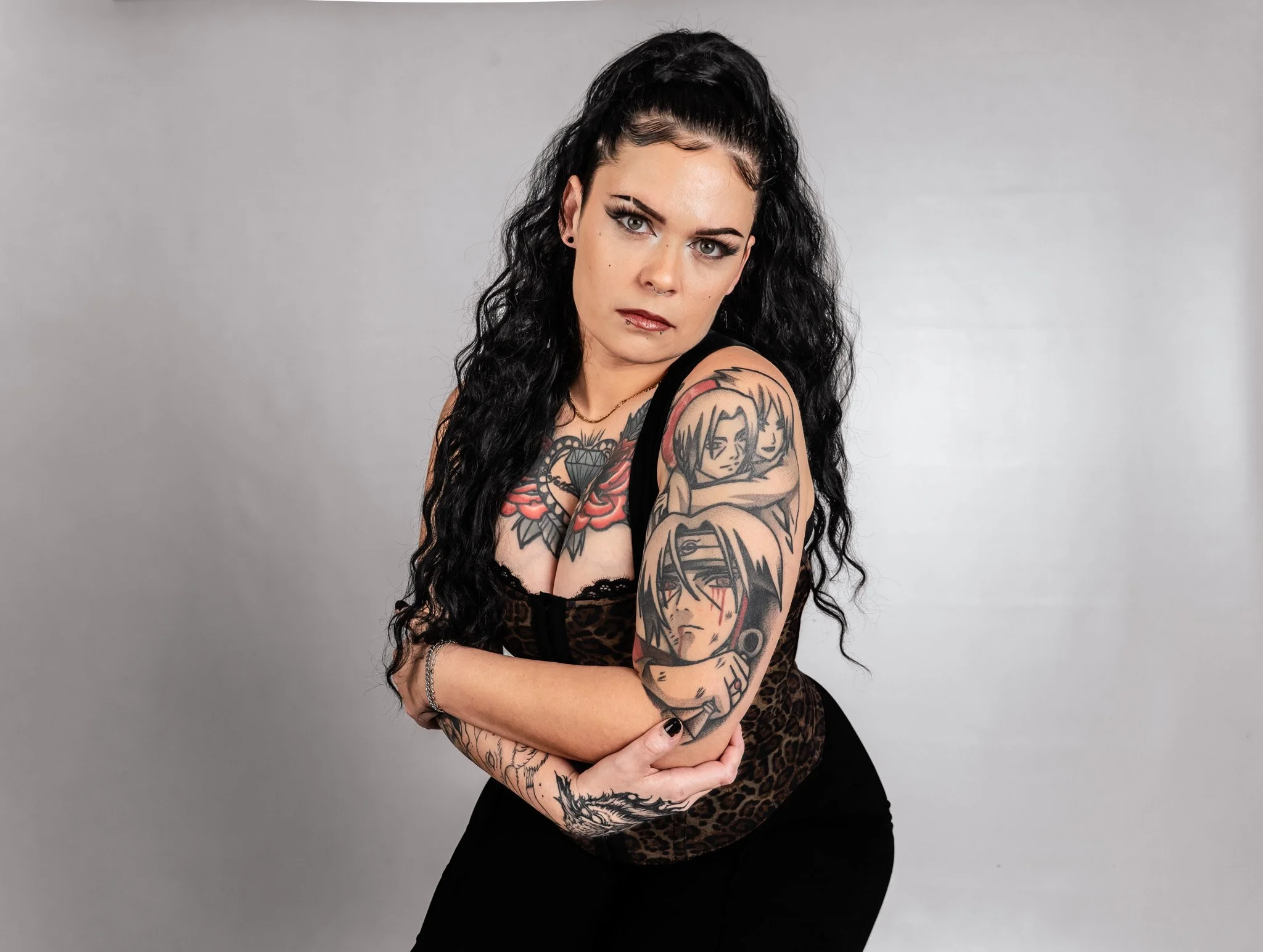 A woman with long black curly hair, tattoos on her arms, wearing a black tank top and black pants, stands with her arms crossed against a plain gray background, looking directly at the camera.