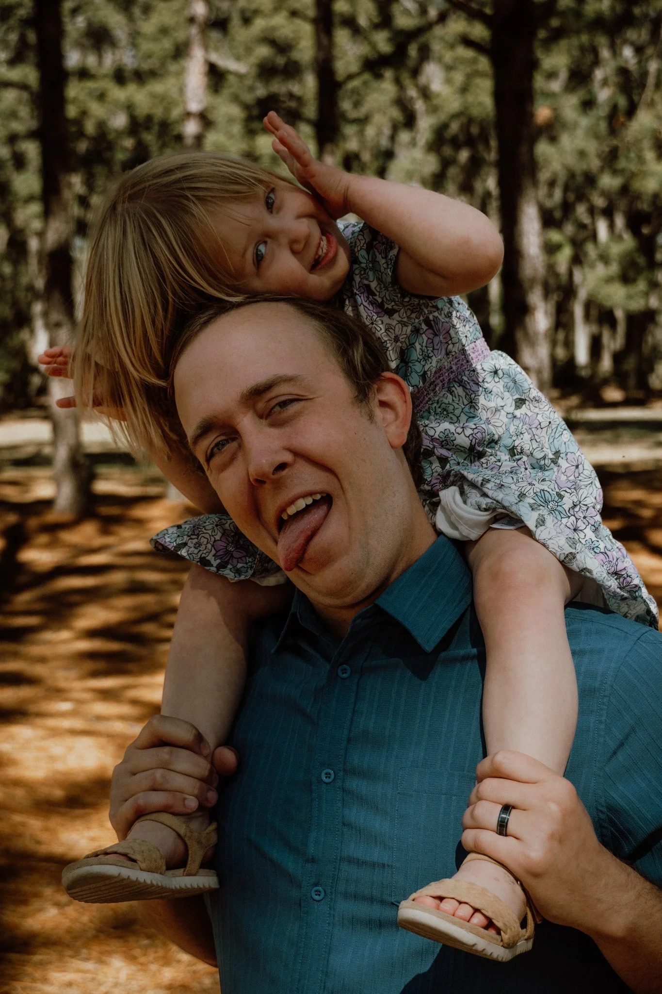 A man with short dark hair carrying a young girl with blond hair on his shoulders in a wooded outdoor setting. The girl is smiling and making a playful gesture by holding her hand near her face. The man is sticking out his tongue and smiling, holding