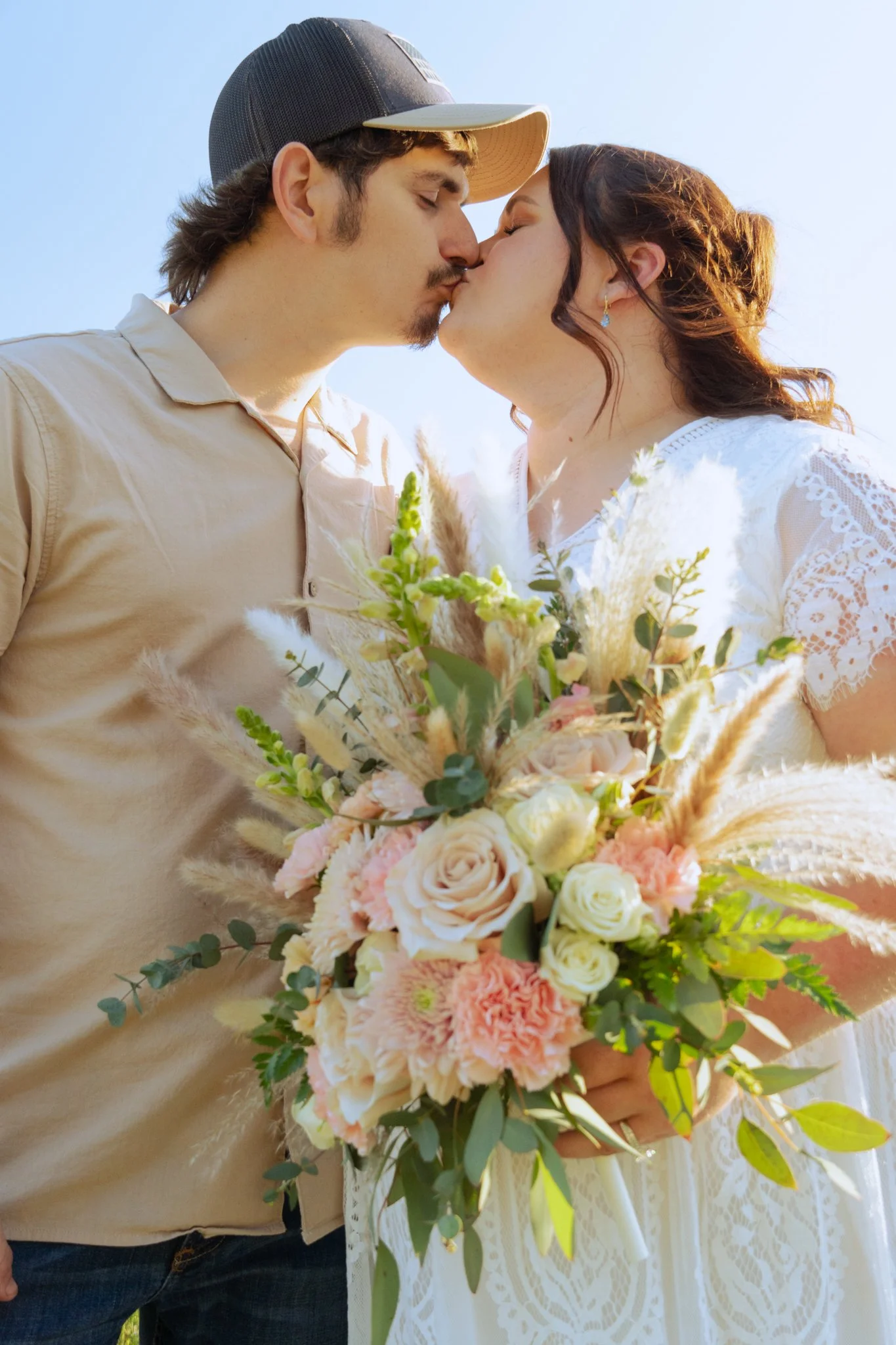 A couple is kissing outdoors under a clear blue sky. The woman is holding a large bouquet of pink and white flowers, wearing a white lace dress, and has dark red hair. The man is wearing a beige shirt, a baseball cap, and has a mustache and beard.