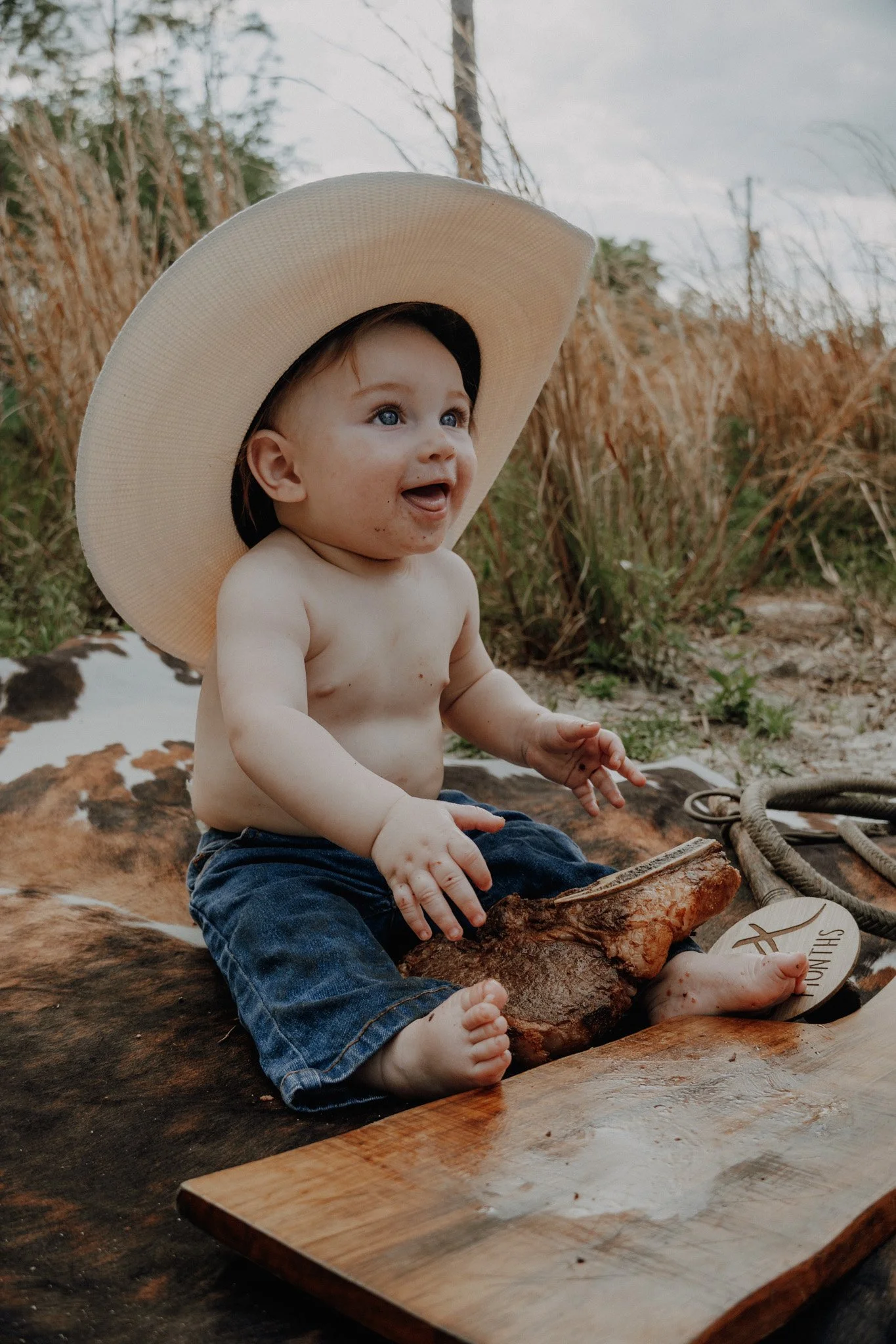 A young boy with blue eyes and light skin, wearing a large straw hat and blue jeans, is sitting on a piece of wood outdoors. He is smiling and looking at something off-camera. There are tall, dried grasses and trees in the background, with a cloudy s