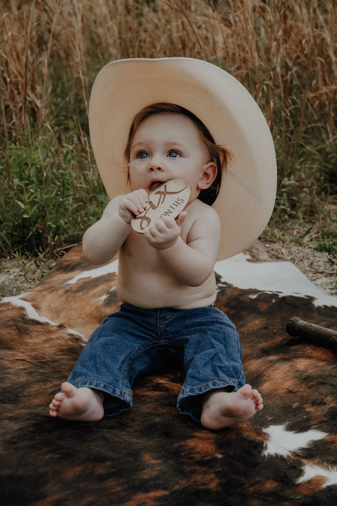 A baby wearing a large beige sunhat, blue jeans, sitting on a cowhide rug outdoors in a field of tall grass, holding a wooden heart-shaped sign with a cross and the word SUNDAY.