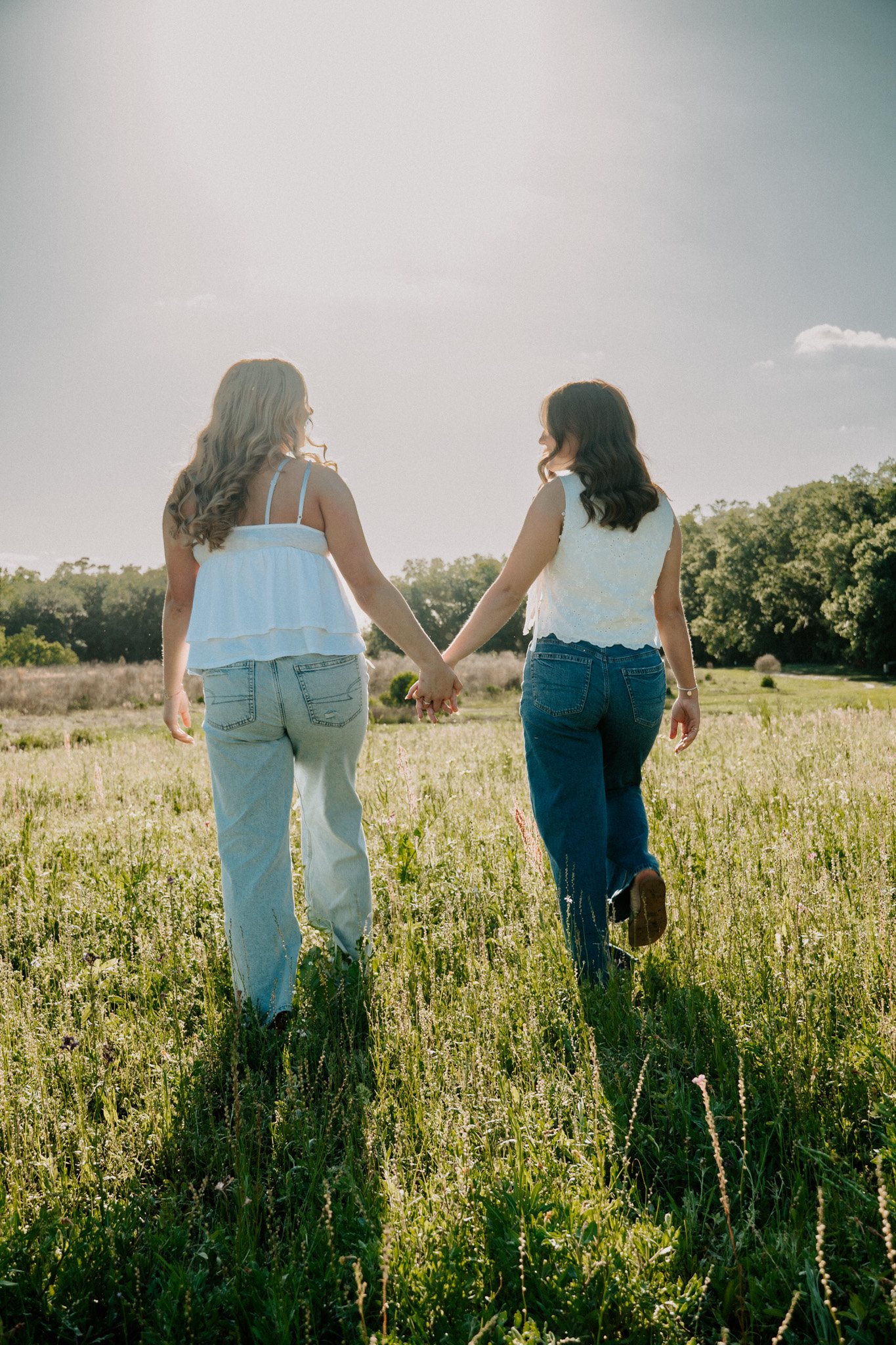 Two women holding hands and walking through a grassy field on a sunny day.