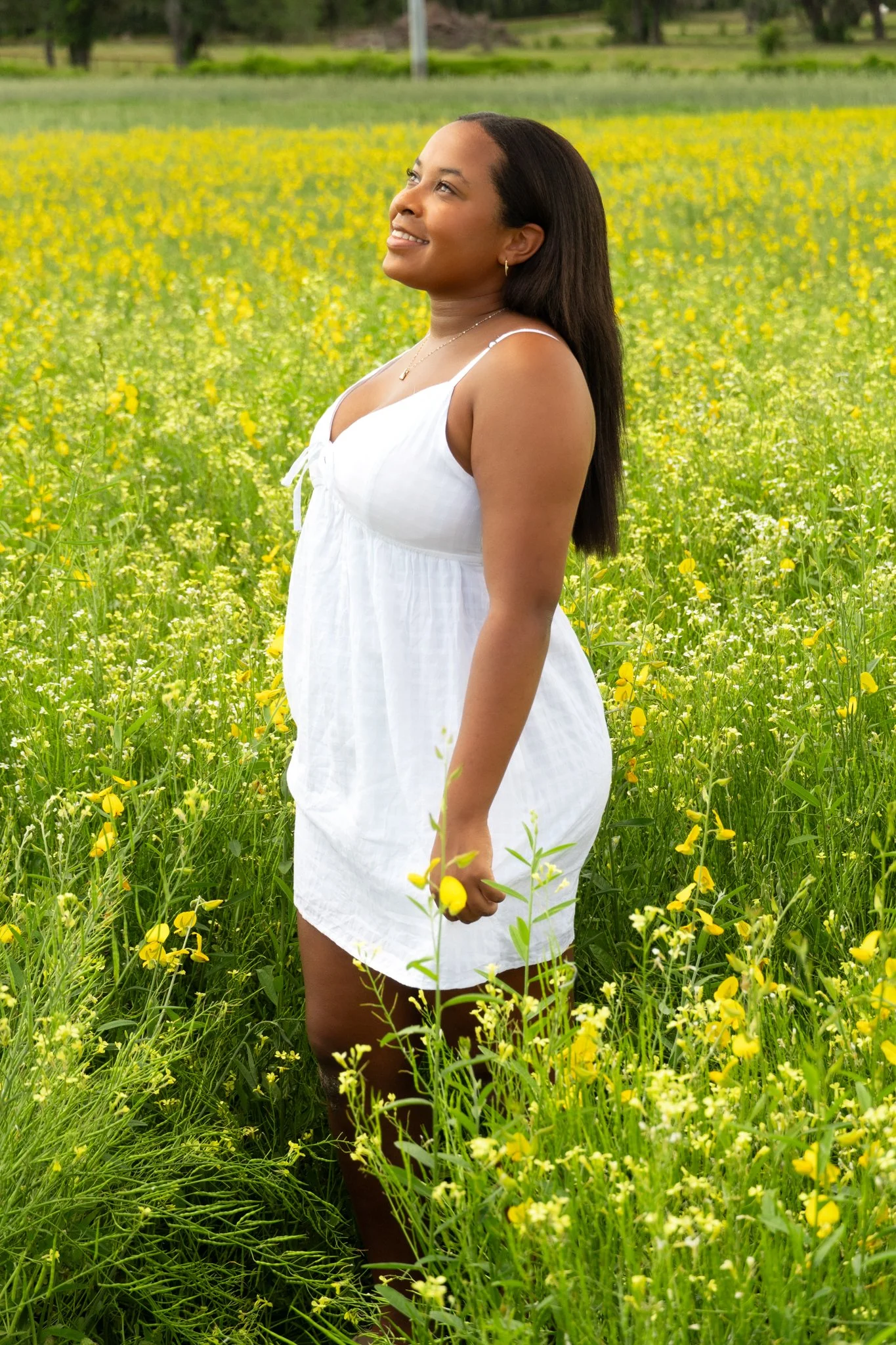 A woman in a white dress standing in a field of yellow wildflowers, smiling and looking to the side.