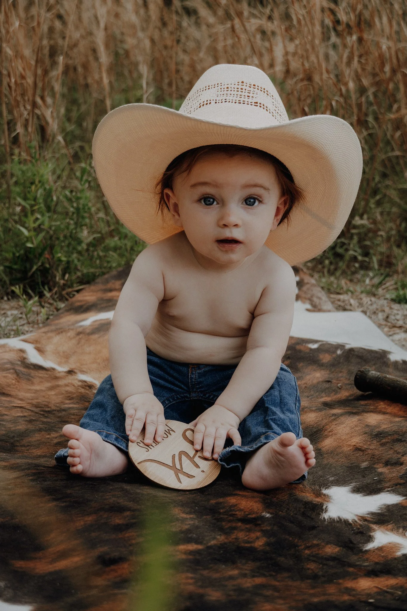 A young boy with blue eyes and brown hair, wearing a large white sunhat and denim shorts, sitting barefoot on a fur rug outdoors near a stream. He has a surprised expression and is holding a small wooden paddle.