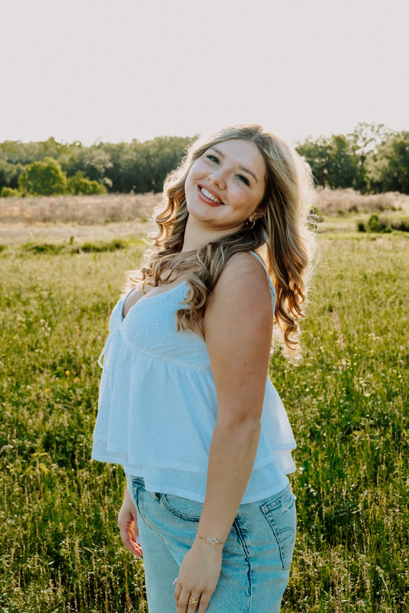 A smiling young woman with wavy blonde hair stands in a grassy field with trees in the background on a sunny day.