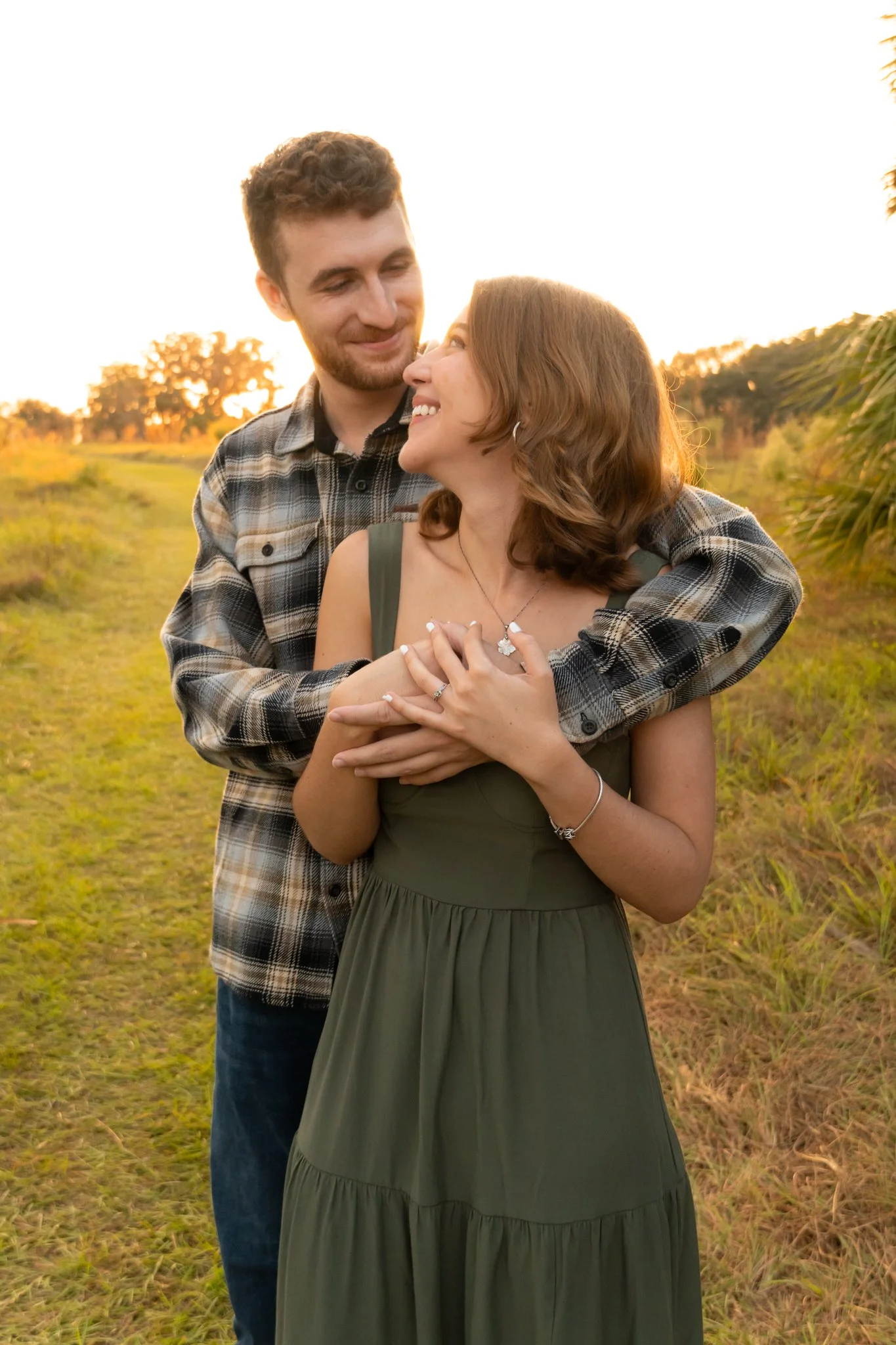 A young couple standing close together outdoors during sunset, with the man smiling at the woman who is smiling back and holding his hands, in a natural setting with trees and grass.