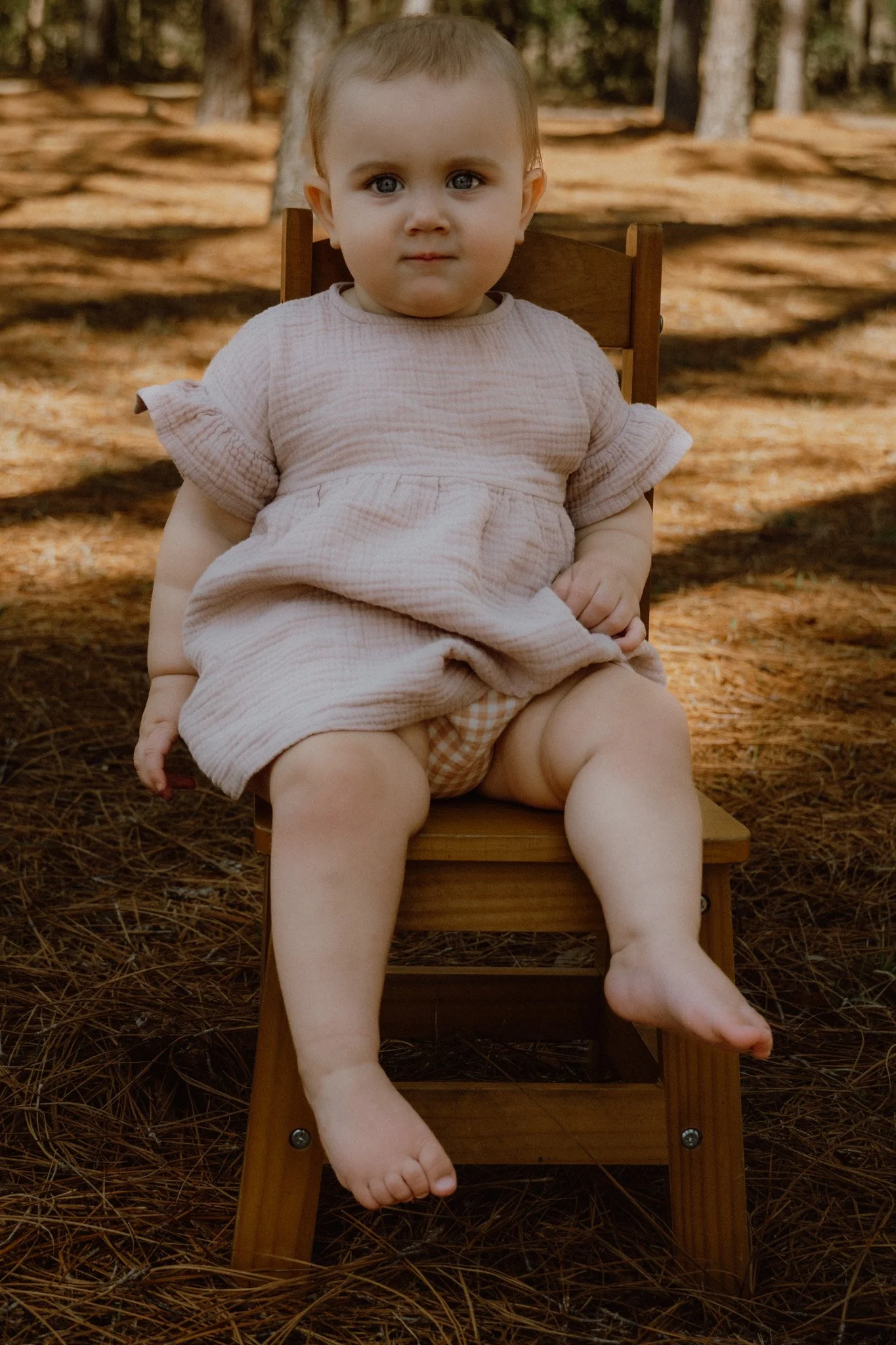 A young child sitting on a wooden chair outdoors with a background of trees and fallen leaves.