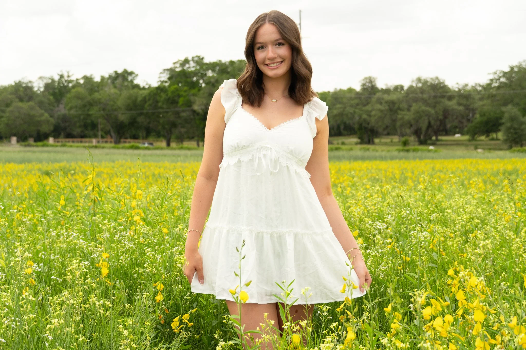 Smiling young woman with brown hair wearing a white dress standing in a yellow flower field with trees and cloudy sky in the background.