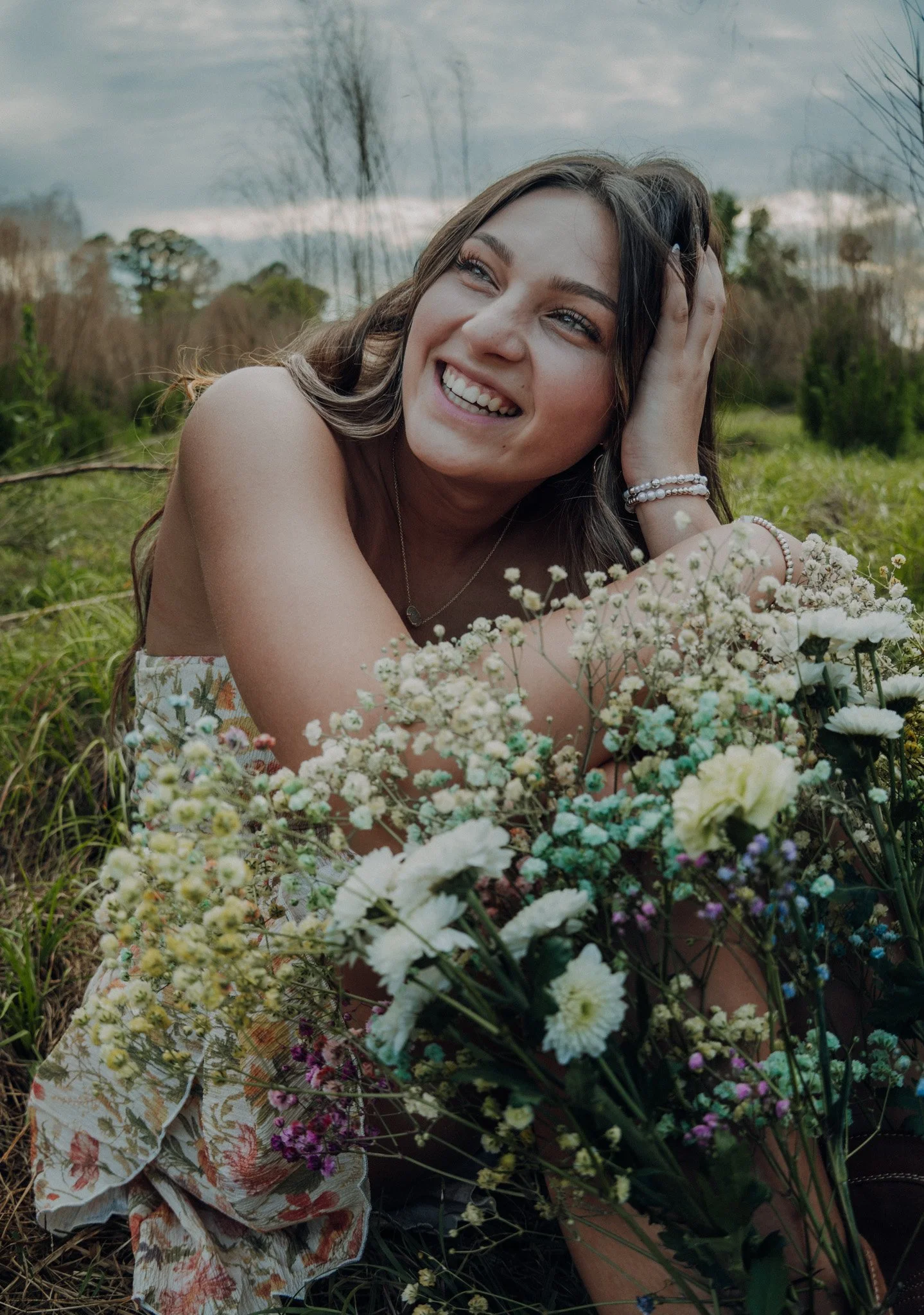 A young woman with long brown hair, smiling, lying on her side in a field of flowers with a cloudy sky and trees in the background.