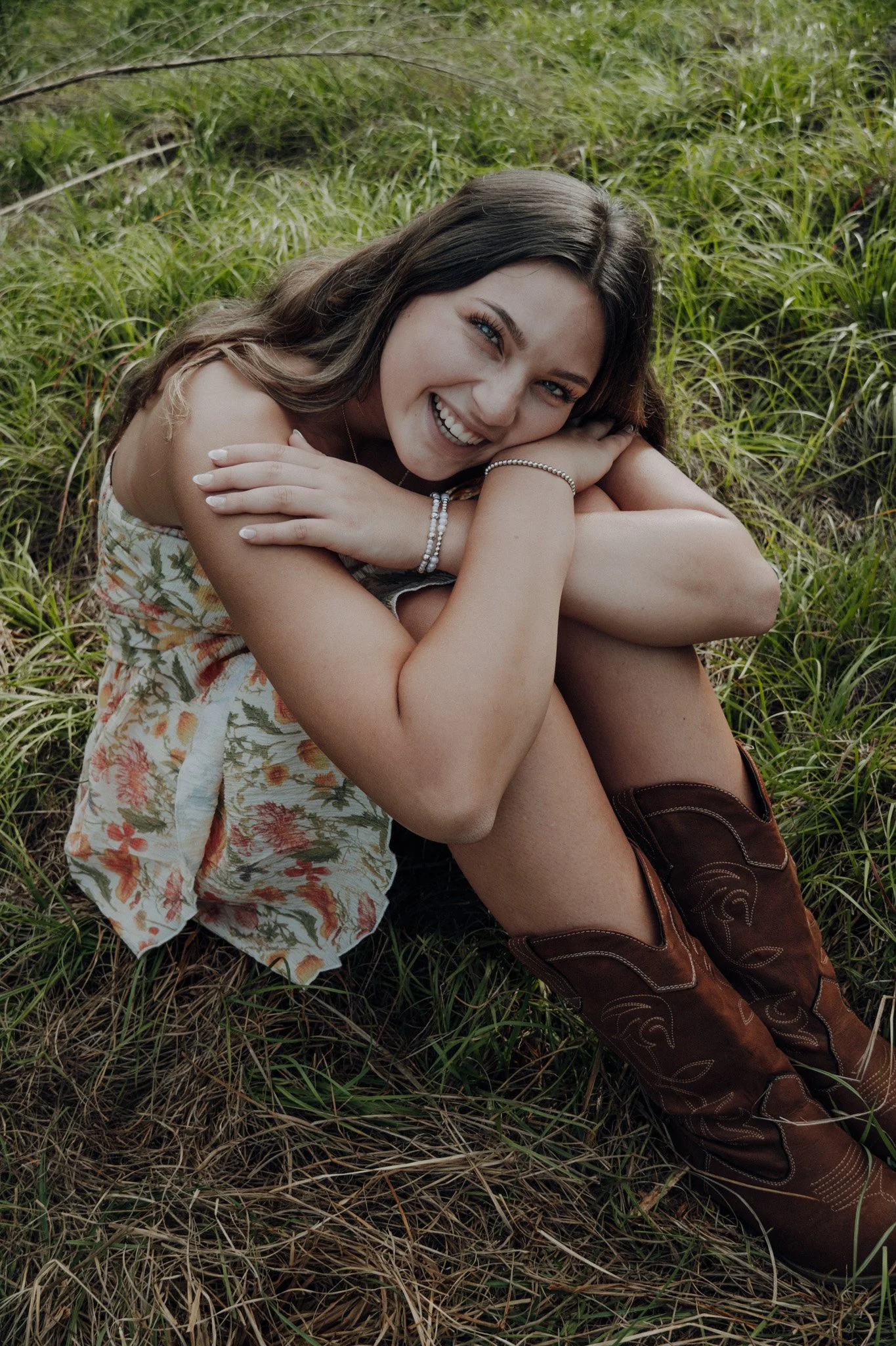 A woman with long brown hair and blue eyes, smiling and lying on her side on green grass, resting her head on her arm, wearing a floral dress, jewelry, and brown cowboy boots.