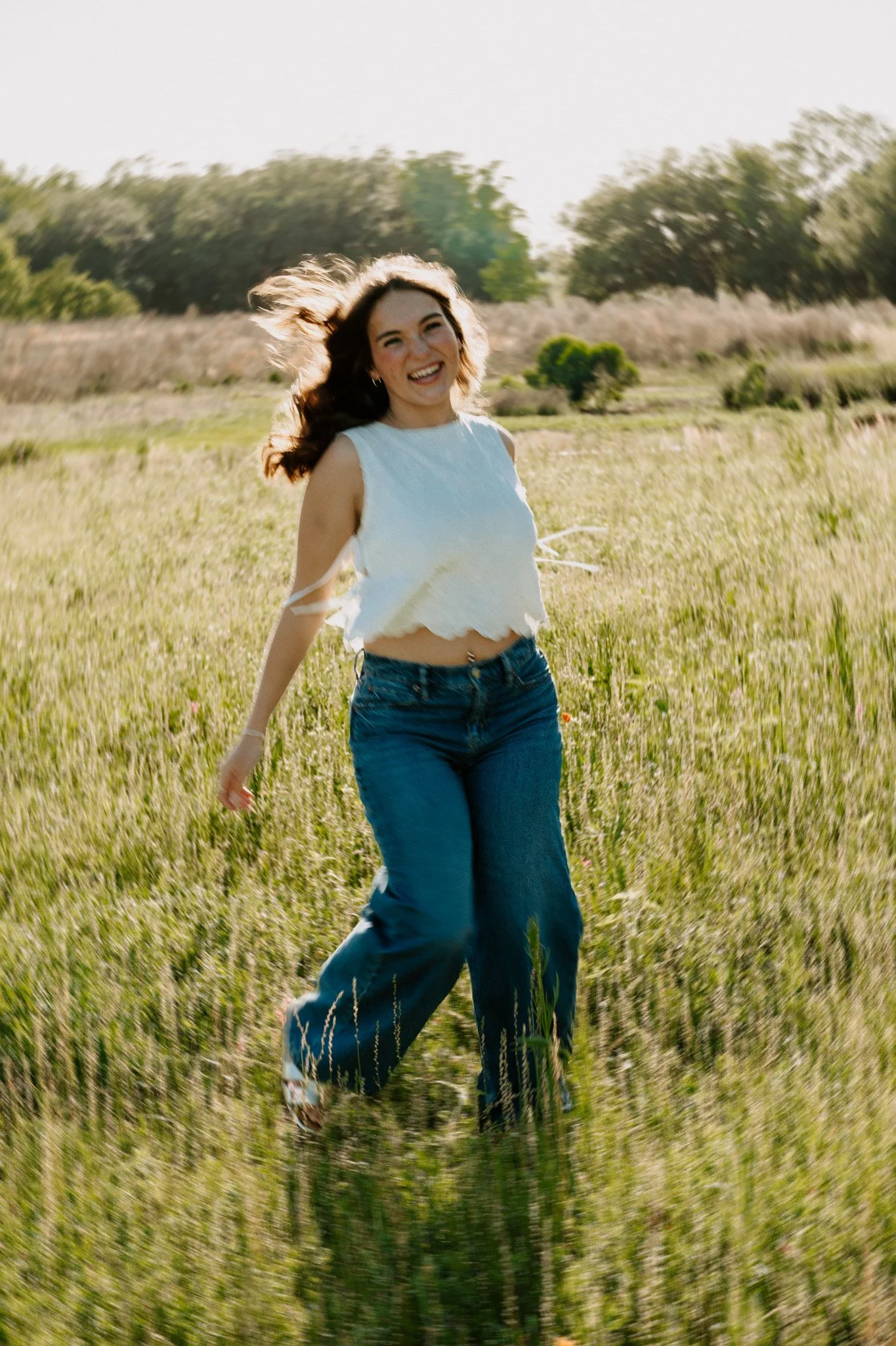 A young woman with dark hair, wearing a white sleeveless top and blue jeans, running through a grassy field with trees in the background, smiling and enjoying the moment.