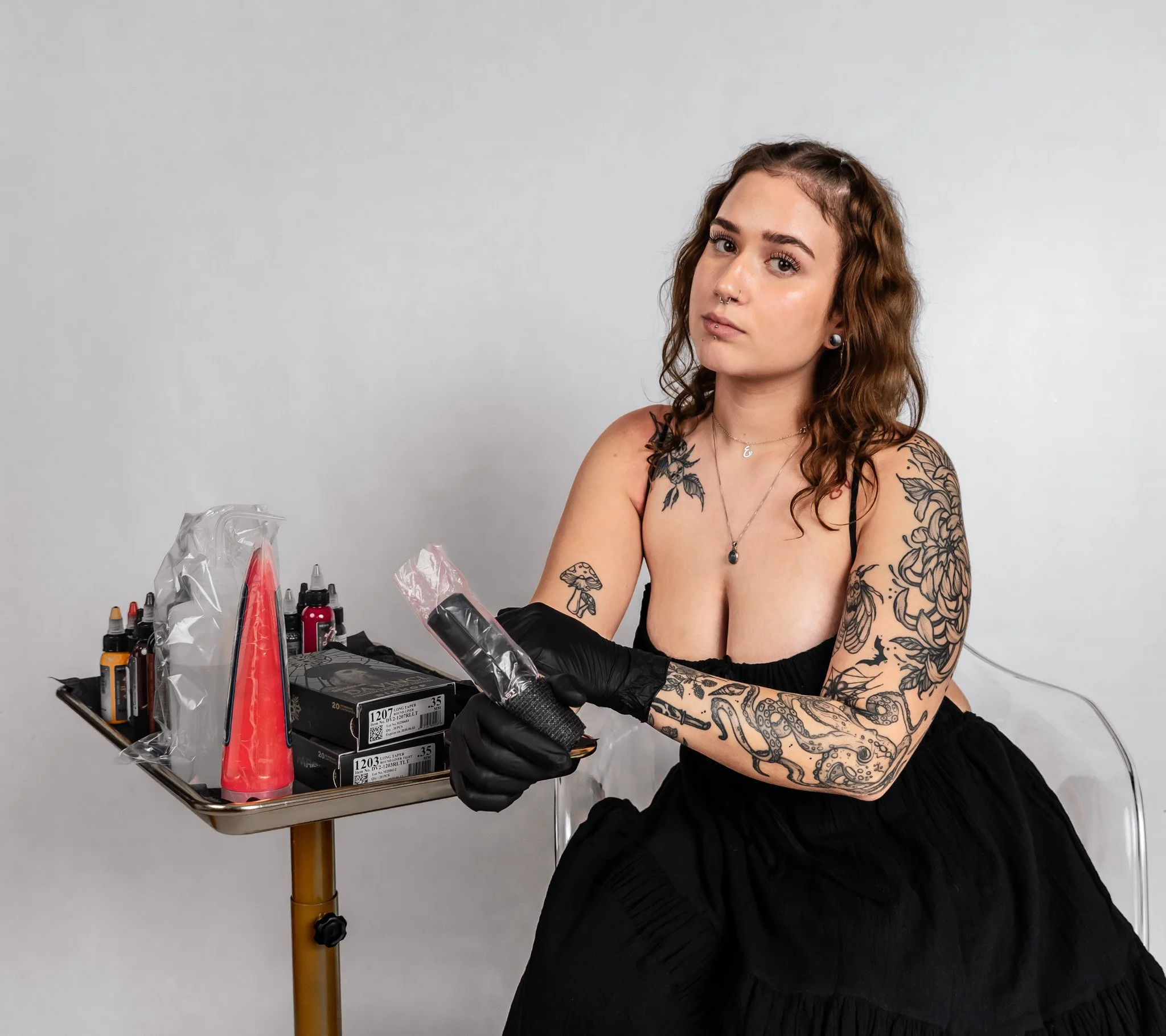 Tattooed woman sitting with a black dress, wearing gloves, holding tattoo supplies, and looking at camera against a plain grey background.
