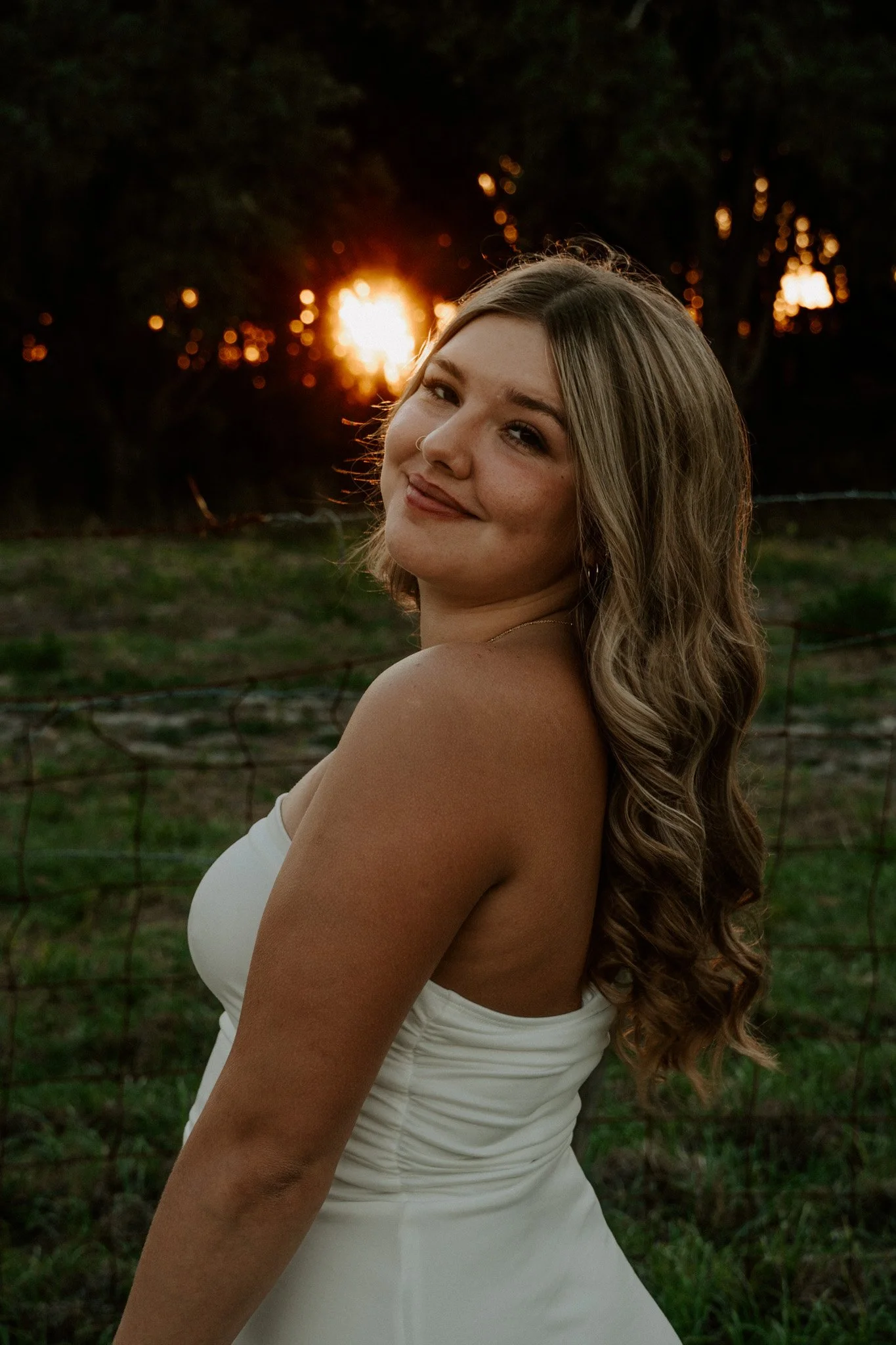 A young woman with long wavy hair and freckles, smiling with closed eyes, standing outdoors at sunset, wearing a white sleeveless dress, with trees and sunlight in the background.
