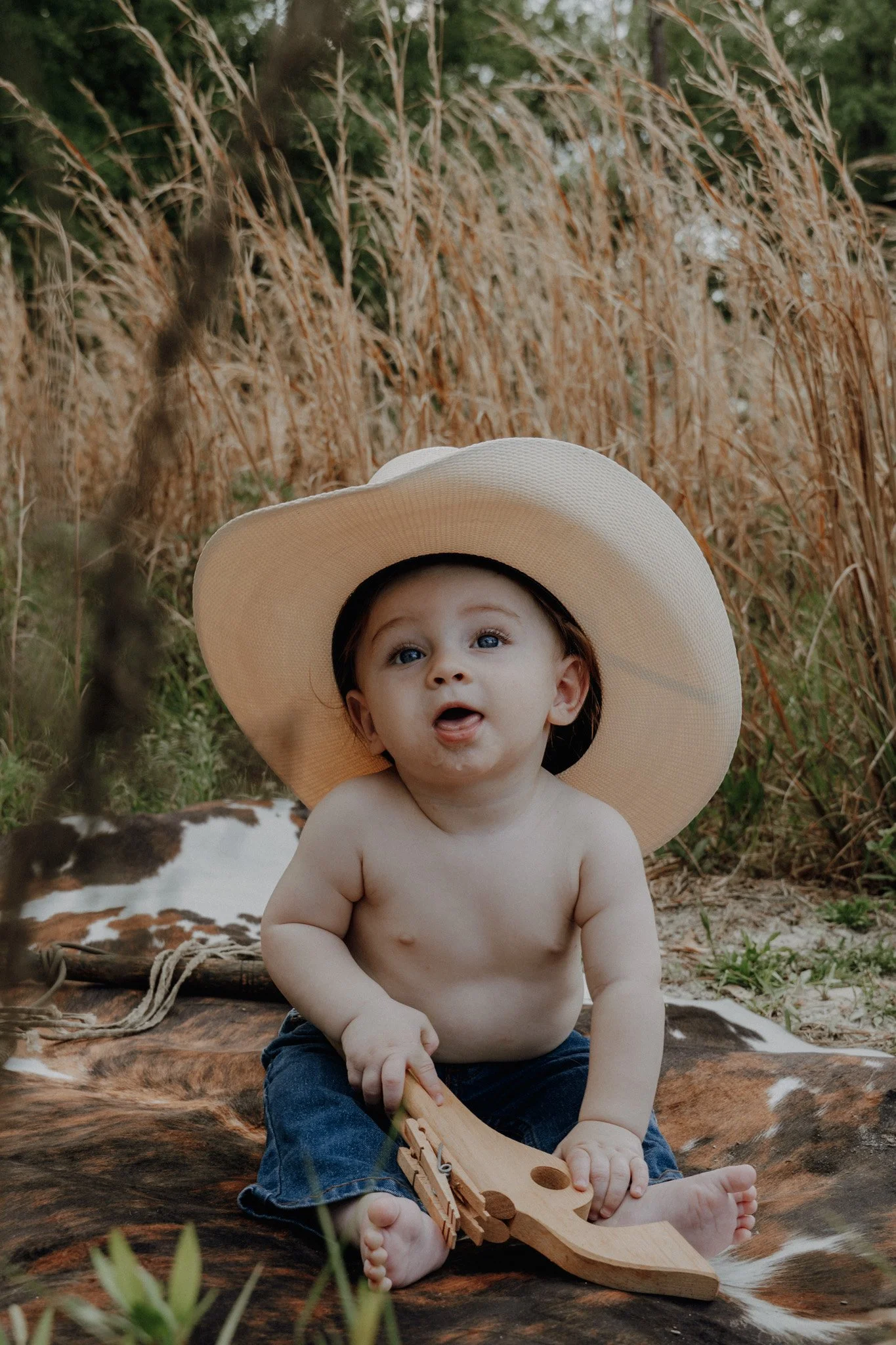 A young child with blue eyes and no shirt, wearing a large straw hat, sits on a cowhide rug outdoors, holding a wooden toy boat. Behind him are tall dry grass and green trees.