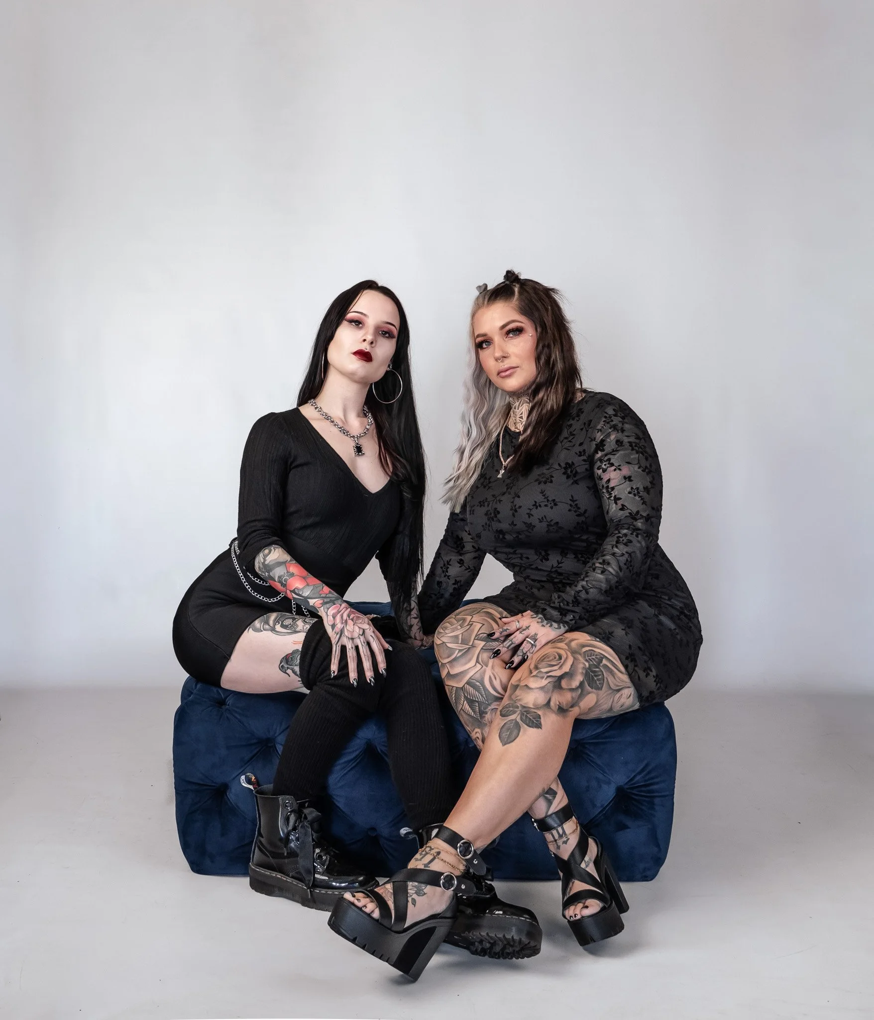 Two women with tattoos sit on a blue cushioned bench against a plain white background. They wear black outfits and platform shoes, with one having dark hair and the other with light gray hair. Both have dramatic makeup and jewelry.