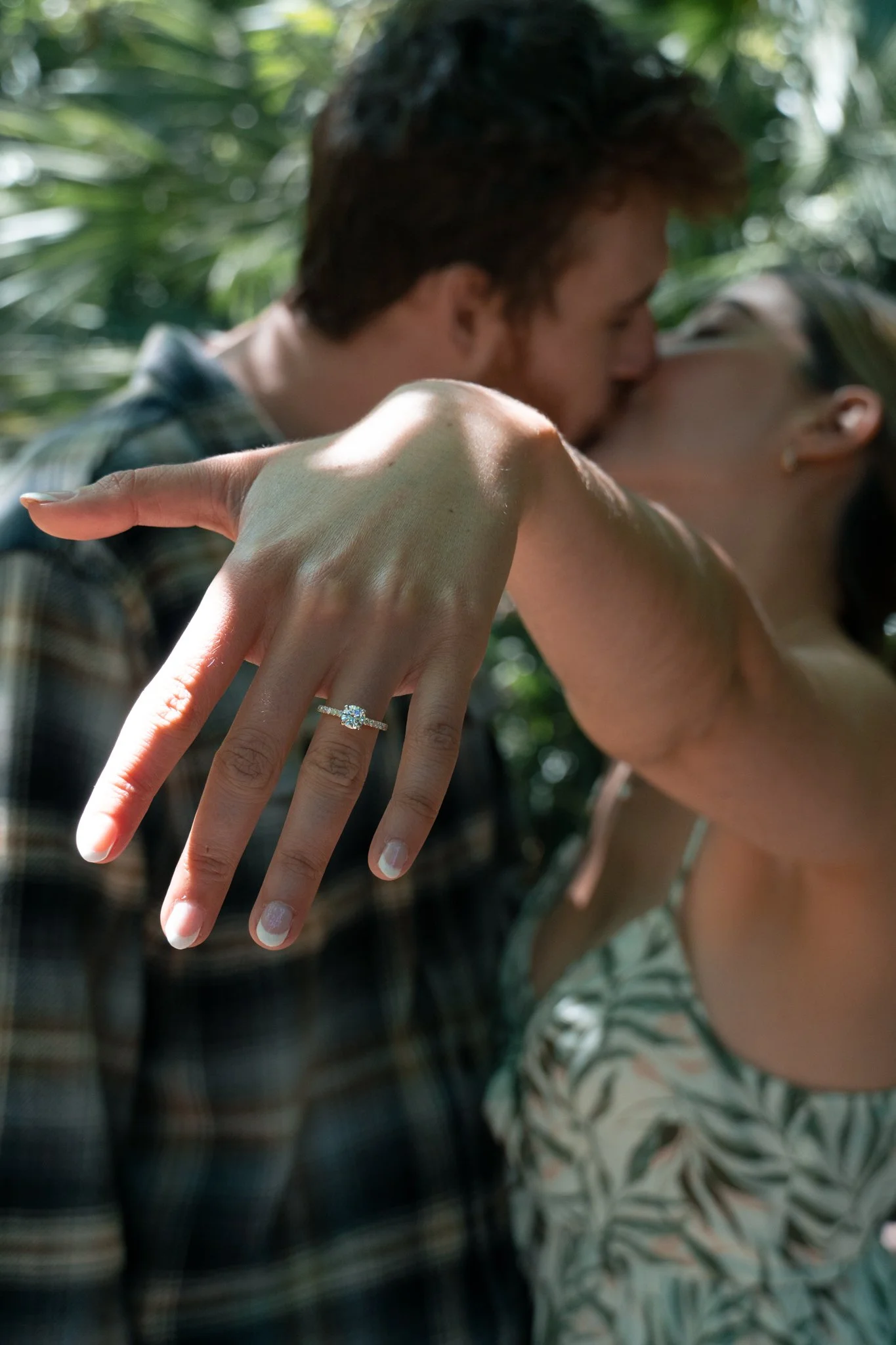 A woman showing her left hand with an engagement ring on her ring finger, while a man kisses her cheek from behind, outdoors with green foliage in the background.