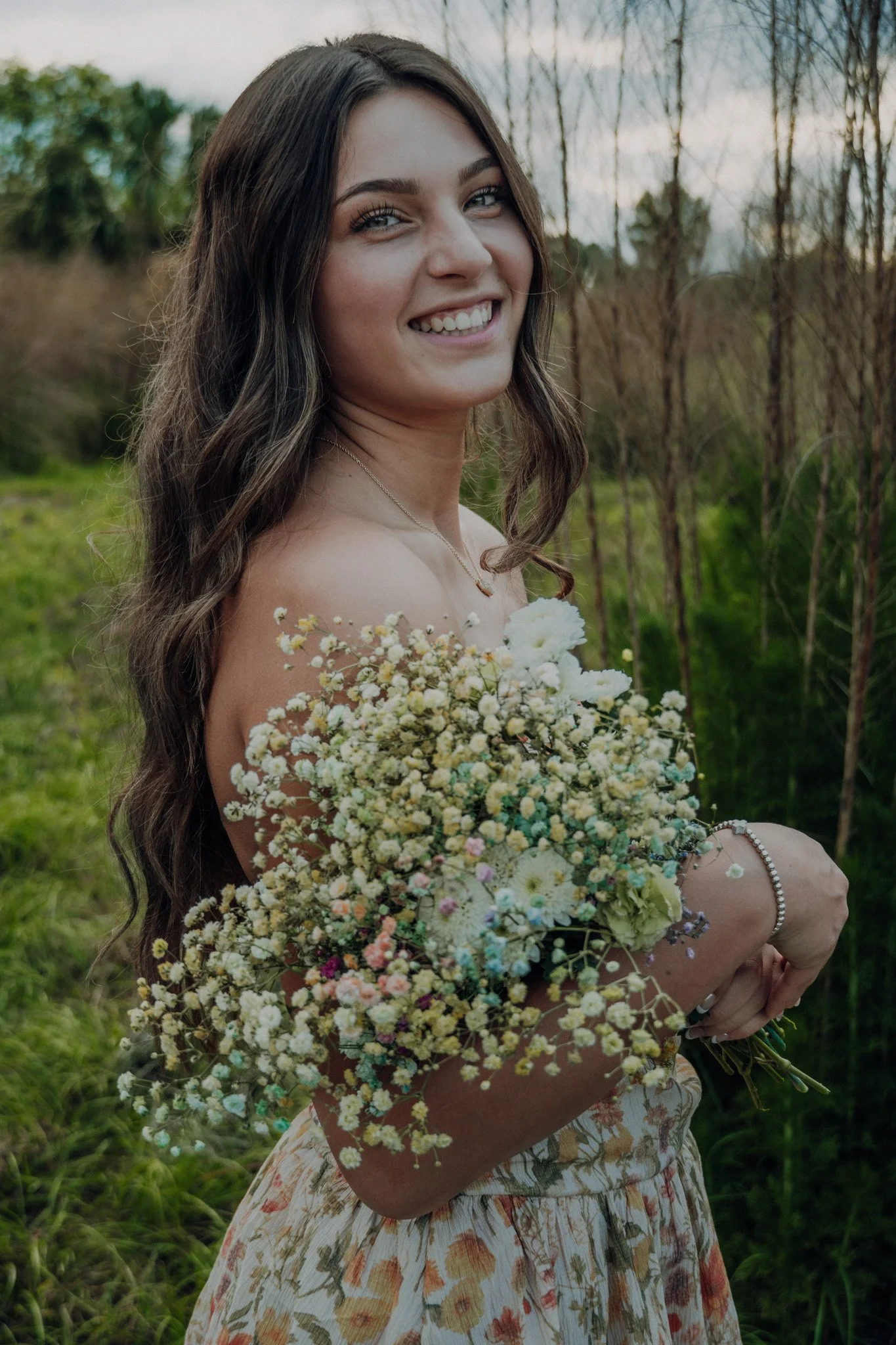Young woman with long wavy brown hair smiling, holding a large bouquet of small white and pastel-colored flowers outdoors during daytime, dressed in a floral dress with a pearl bracelet and necklace, in a natural setting with green grass and plants i