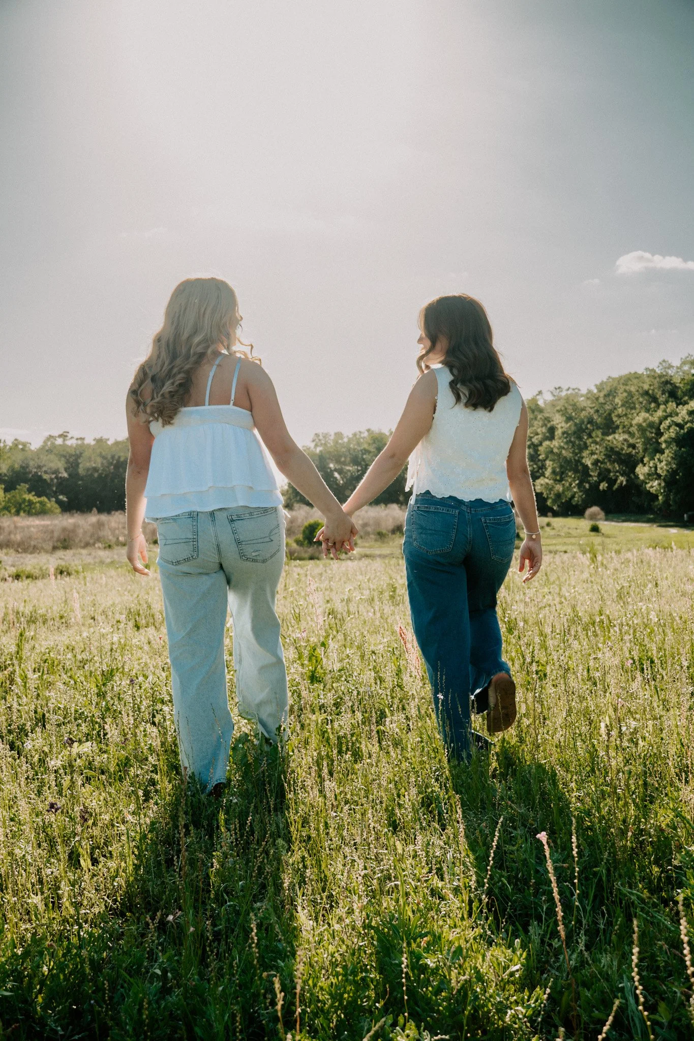Two women holding hands and walking through a grassy field on a sunny day, with trees in the background.