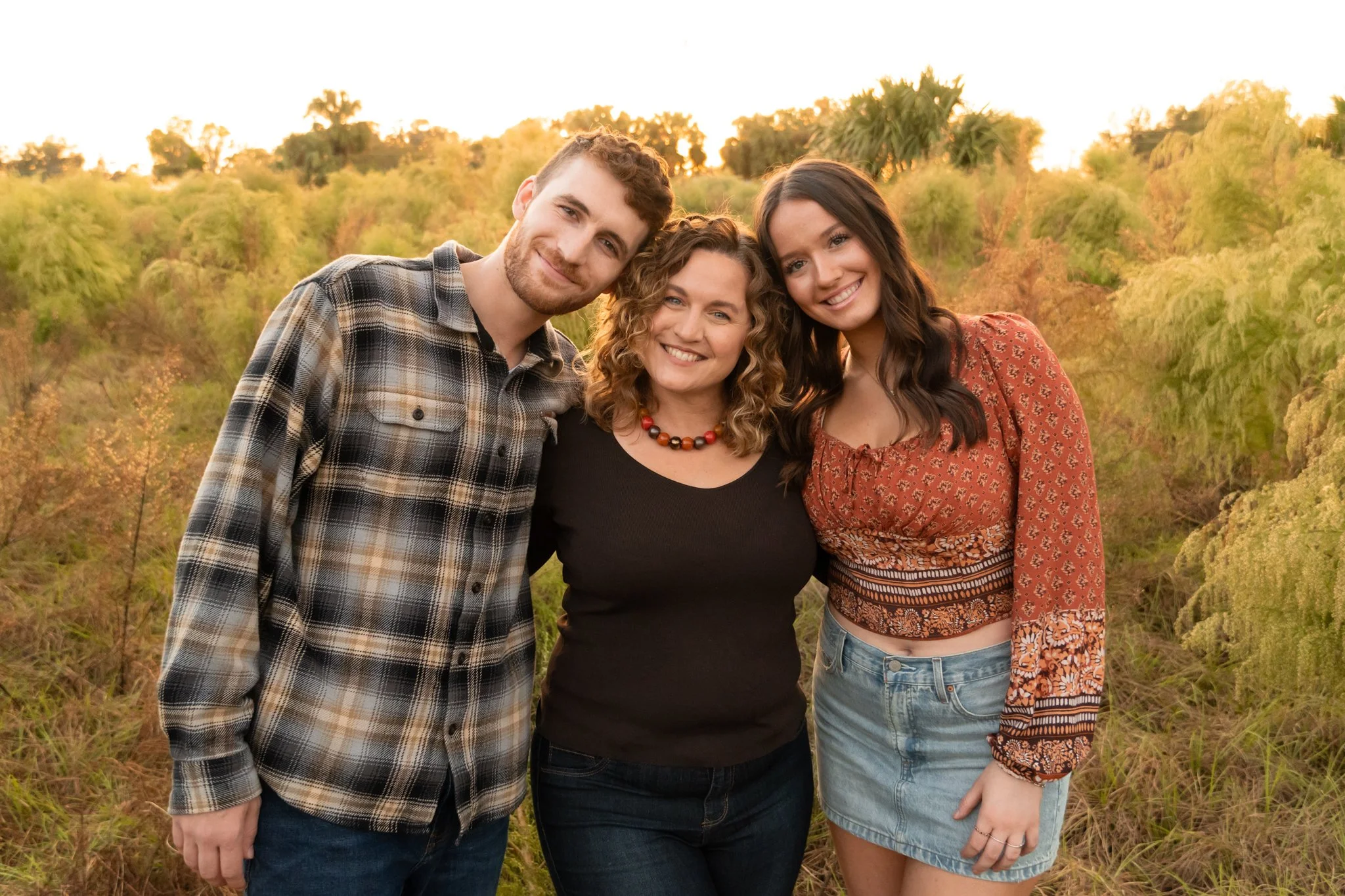 Three smiling women and a man posing outdoors in front of trees at sunset.