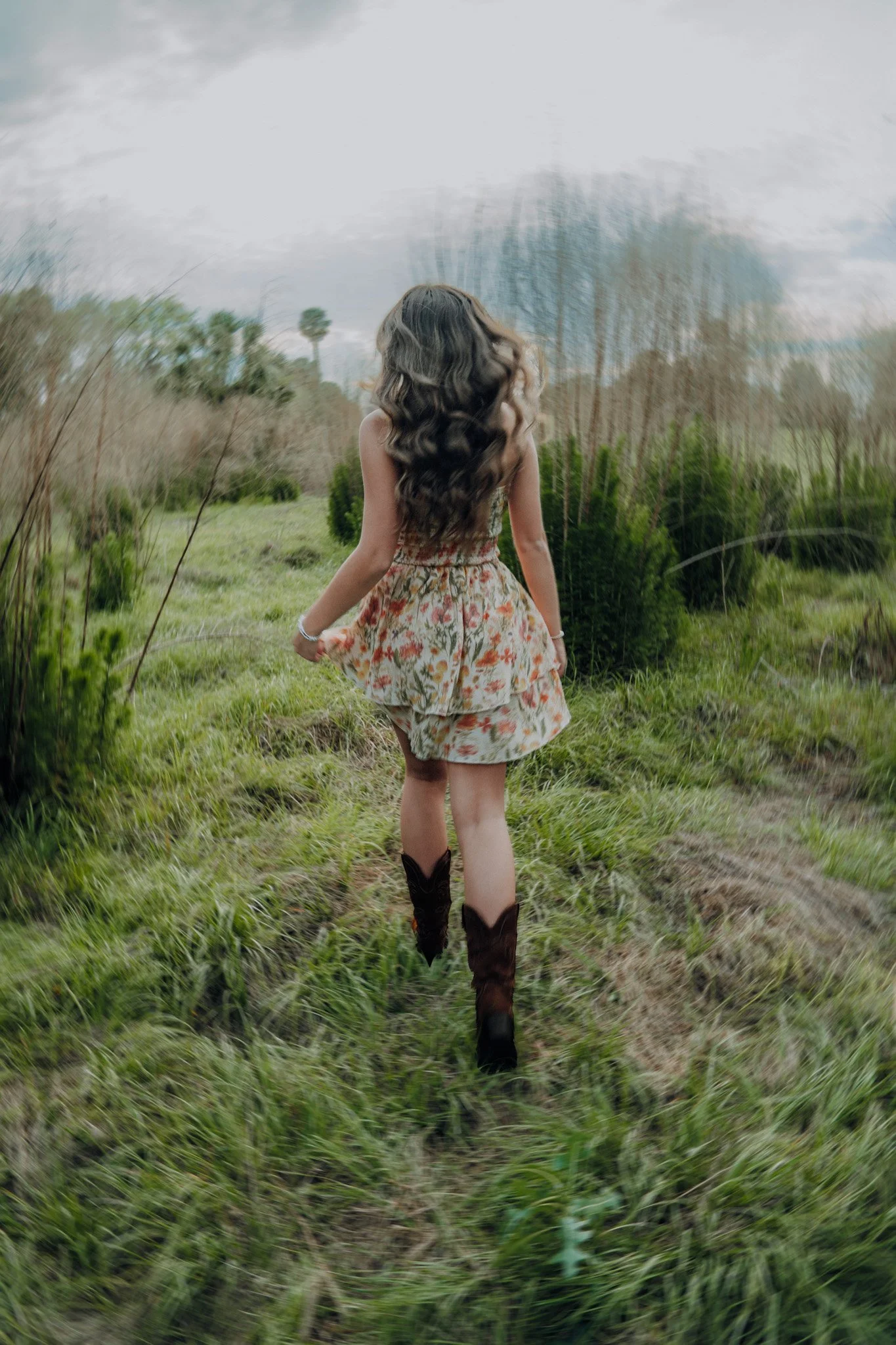 A girl walking on a dirt path through a grassy field, wearing a floral dress and cowboy boots, with trees and cloudy sky in the background.