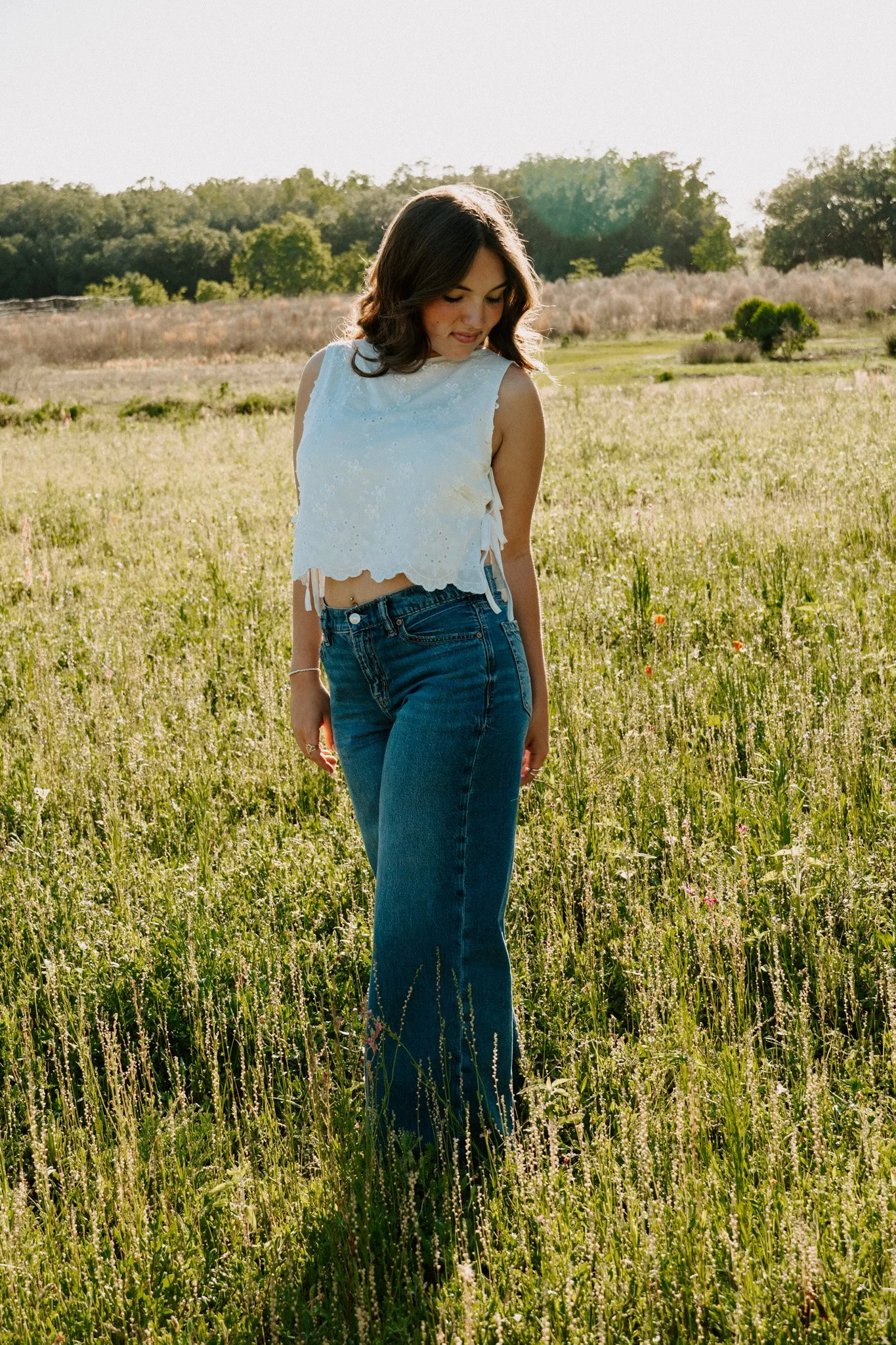 A young woman with dark, wavy hair stands in a grassy field during sunset, wearing a white sleeveless top and blue jeans, looking down and smiling.