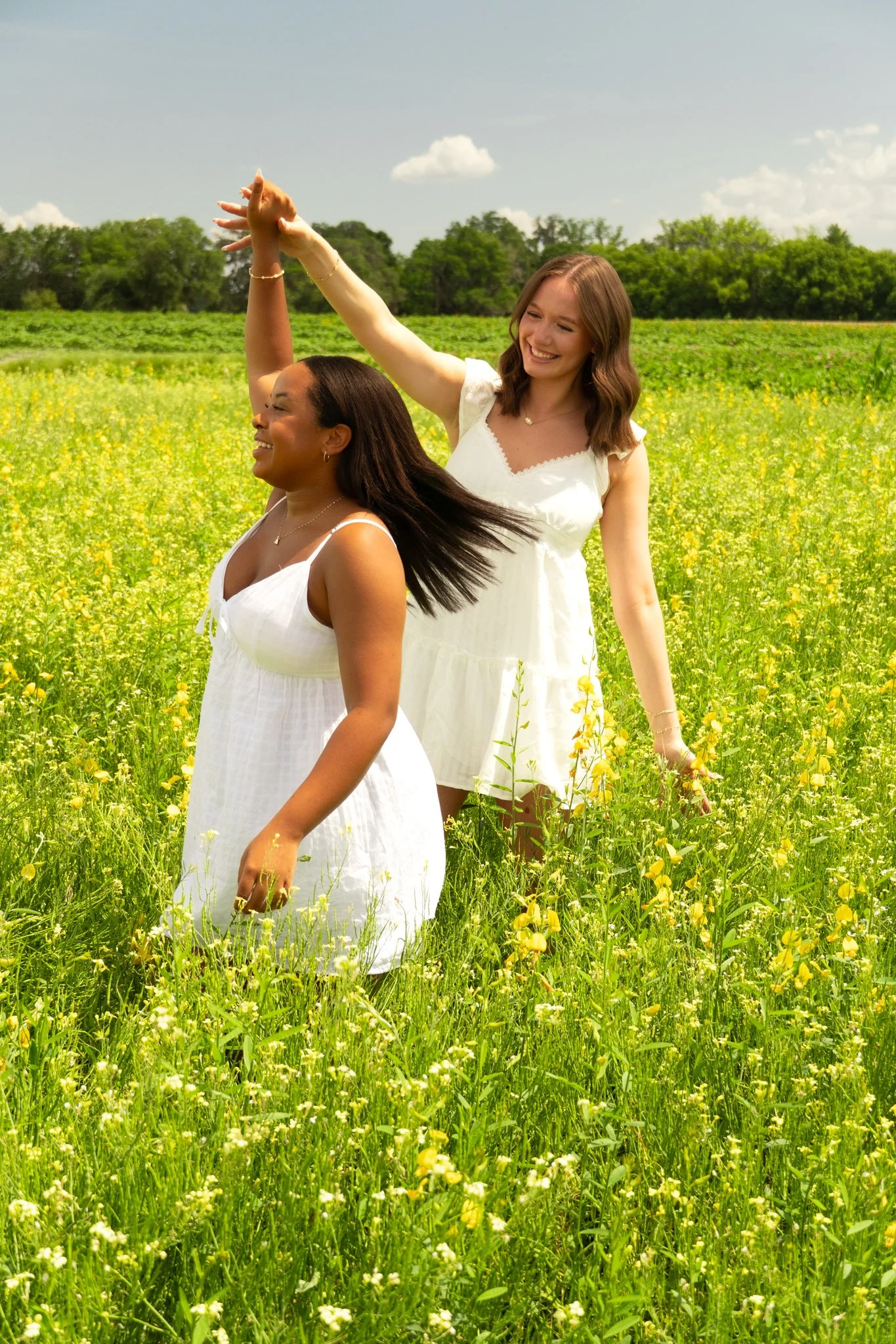 Two women in white dresses dancing and smiling in a green field of yellow flowers with trees and a partly cloudy sky in the background.