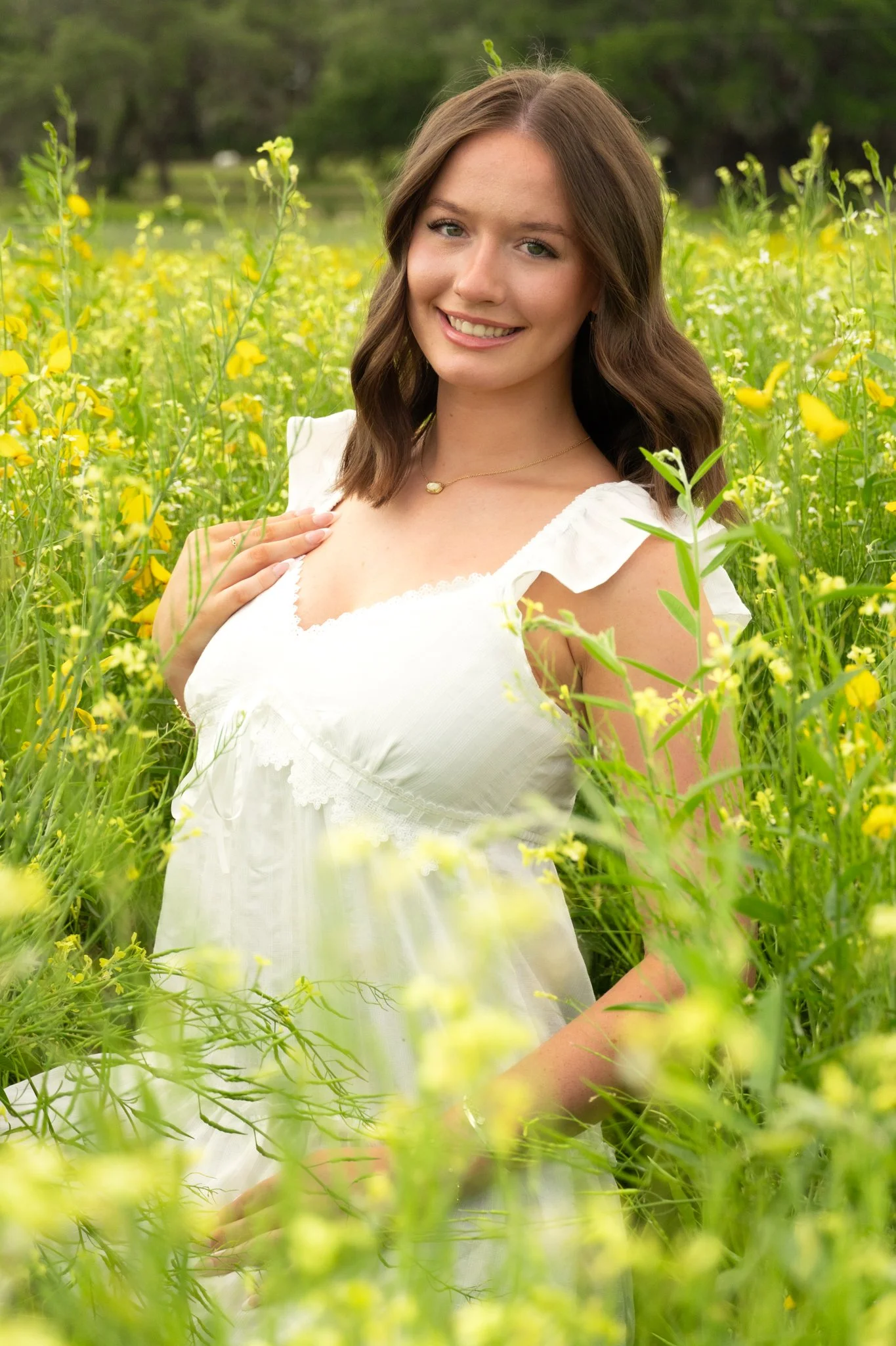 A young woman with brown hair wearing a white dress and a gold necklace standing in a field of yellow flowers, smiling at the camera.