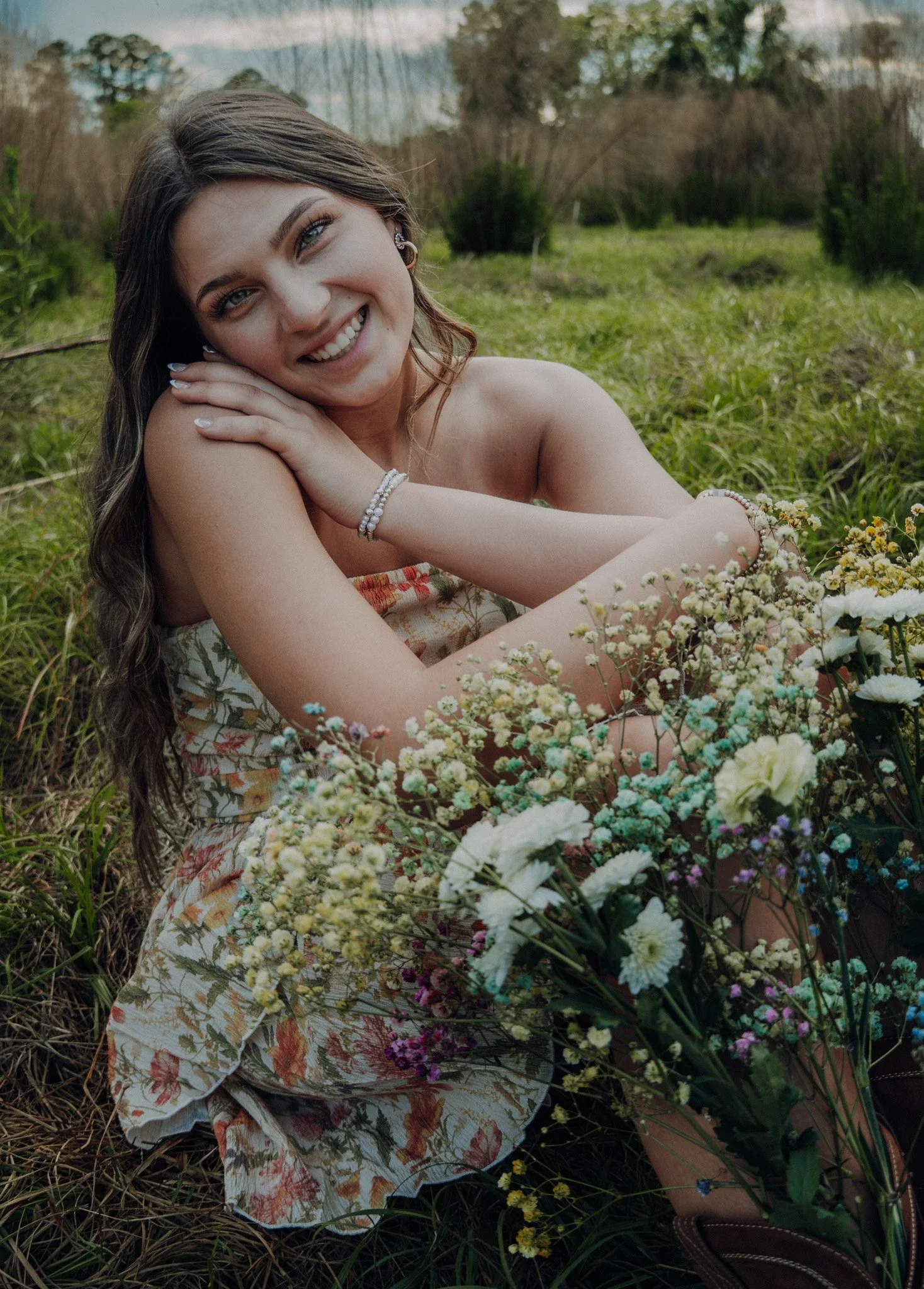 A young woman with long brown hair and blue eyes lying in a grassy field, smiling at the camera, surrounded by flowers and greenery.