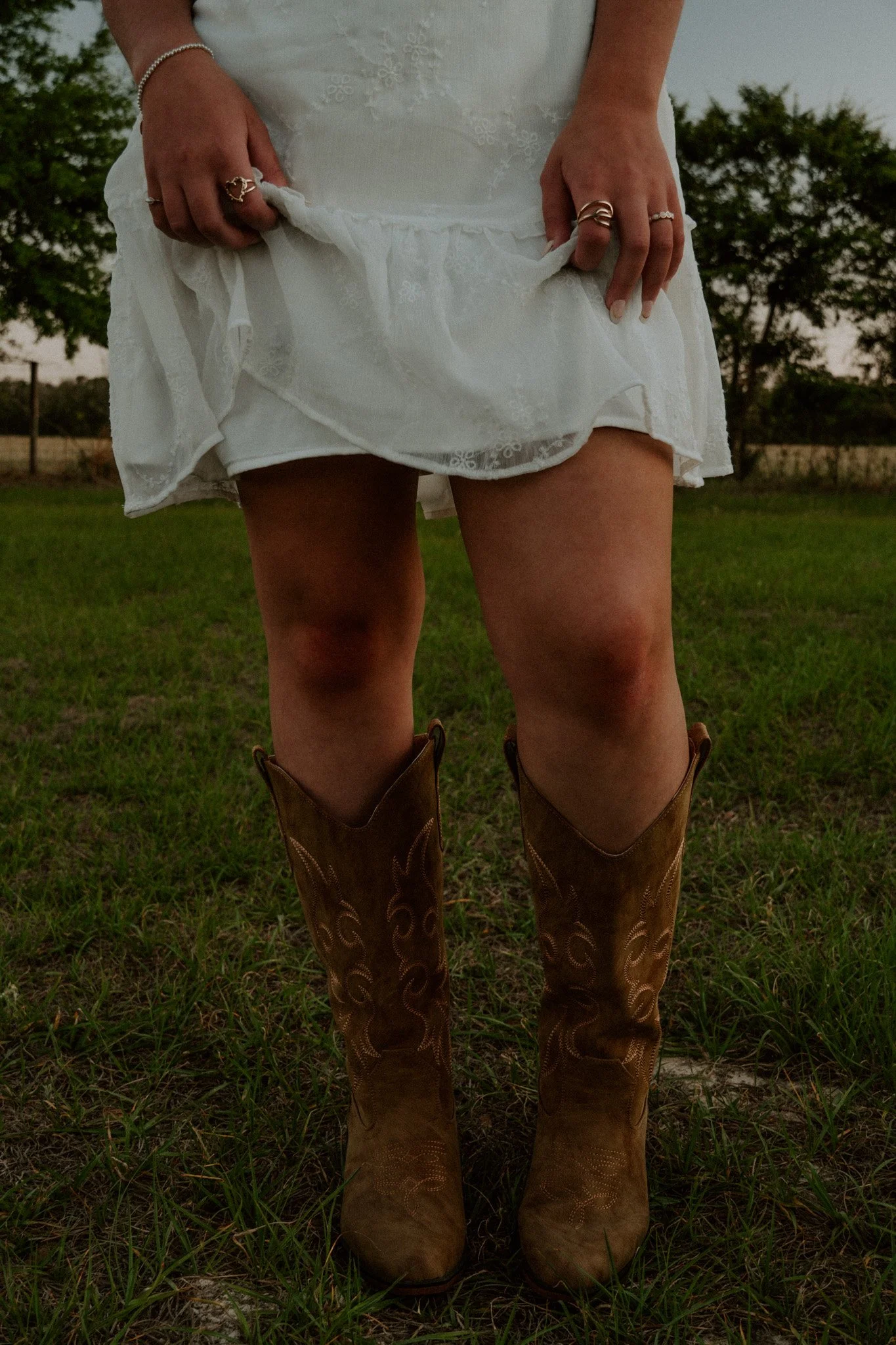A person wearing a white dress, holding up the hem to reveal the underside, and brown cowboy boots standing on grass, with trees and a fence in the background during dusk.