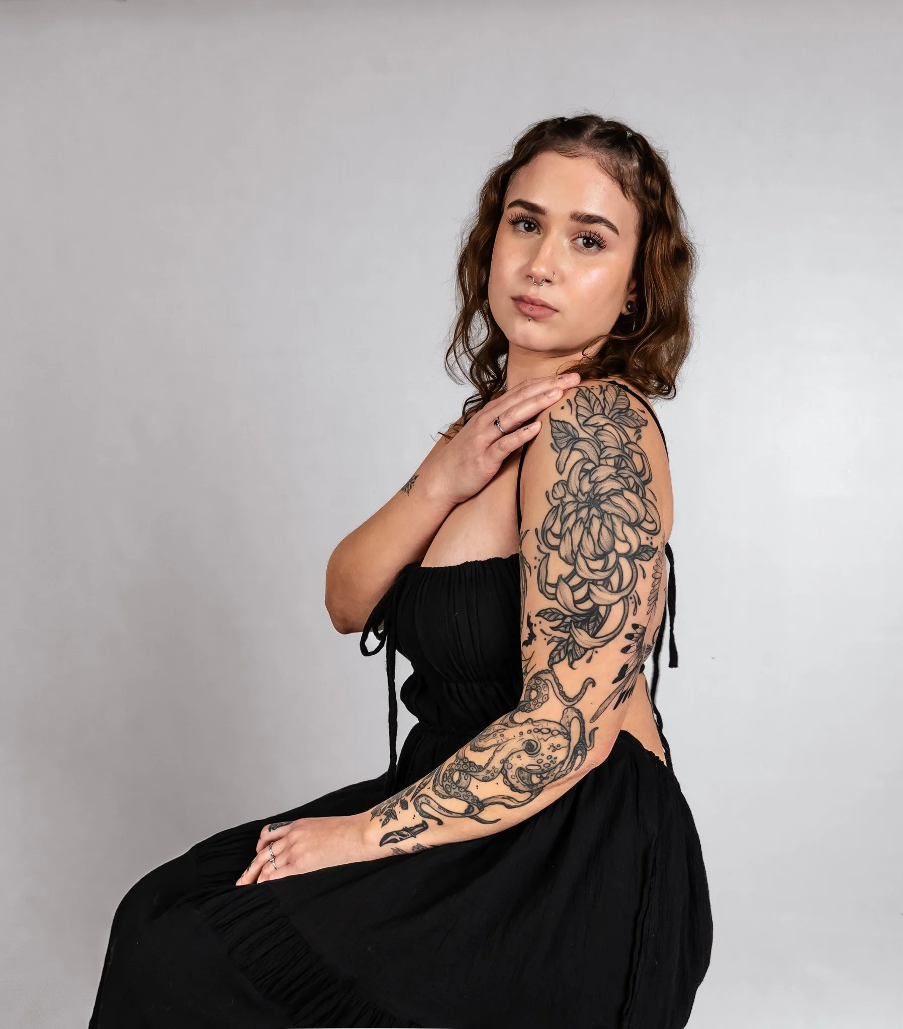 A woman with brown hair and tattoos on her arm, wearing a black sleeveless dress, posing against a plain gray background.
