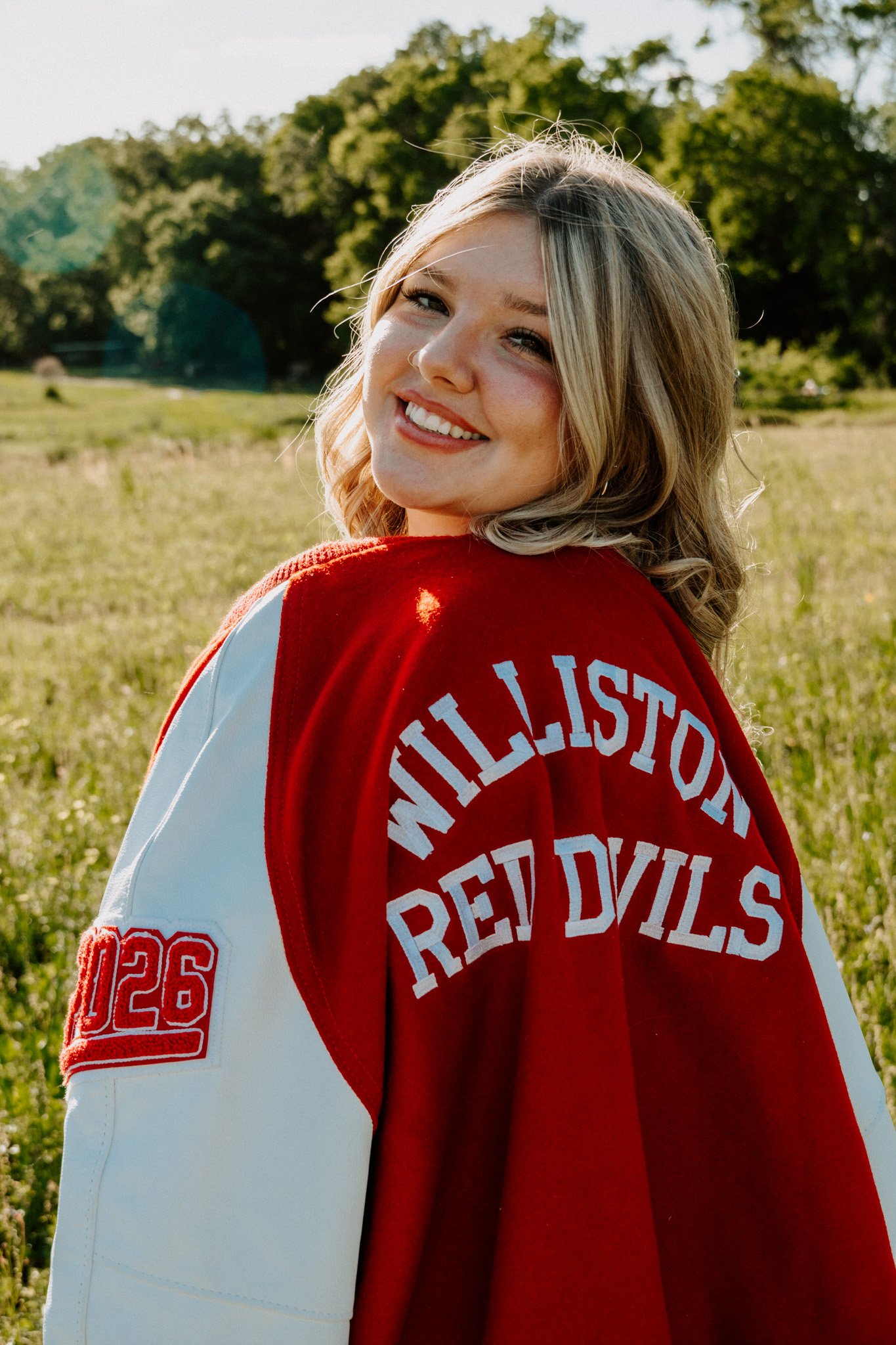 A young woman with blonde hair smiling, wearing a red and white hockey jersey, standing outdoors in a grassy area with trees in the background.