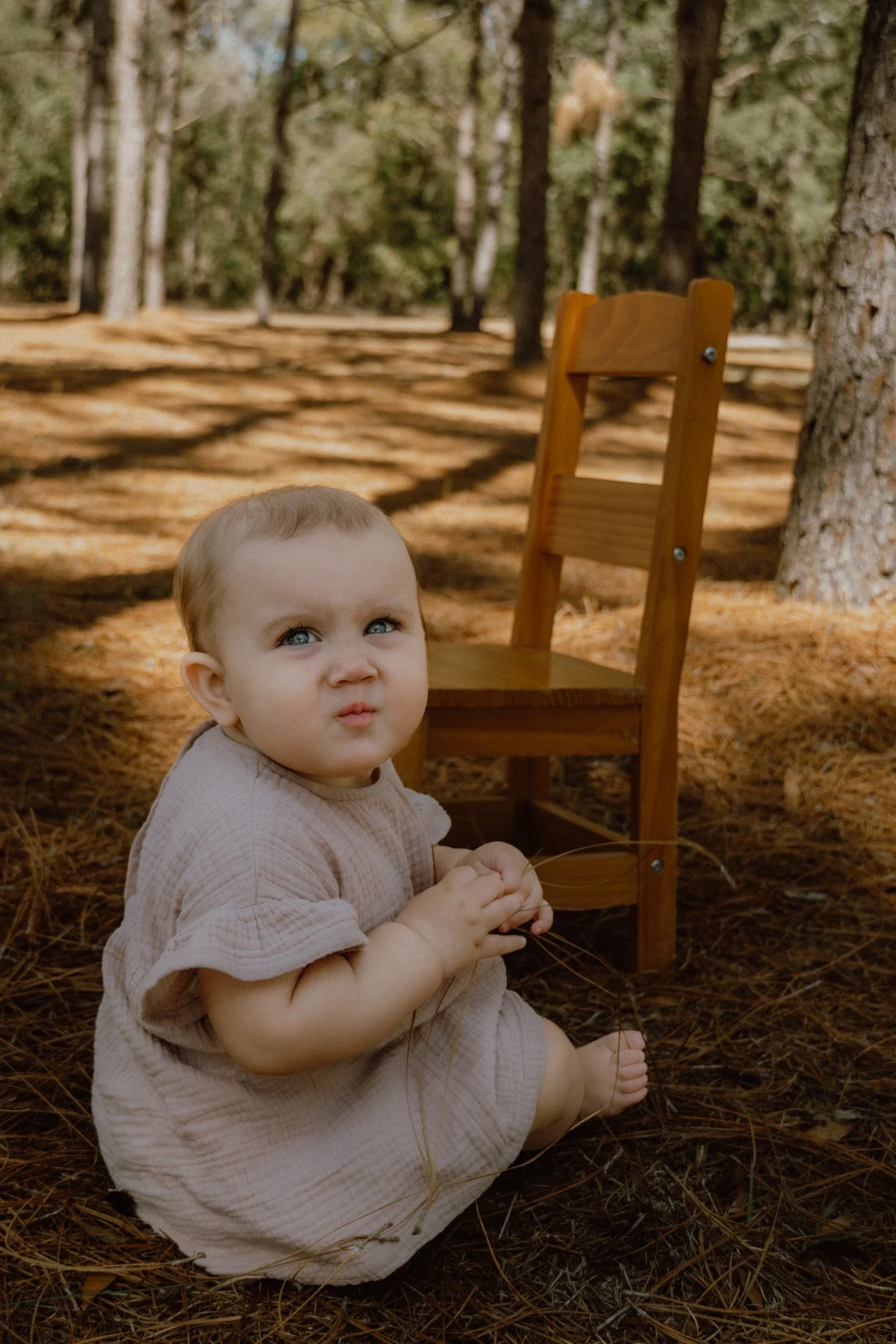 A young child with blue eyes and light hair sitting on the ground in a forest, holding a small branch. There is a wooden chair behind the child, and the forest has tall trees and a carpet of pine needles.
