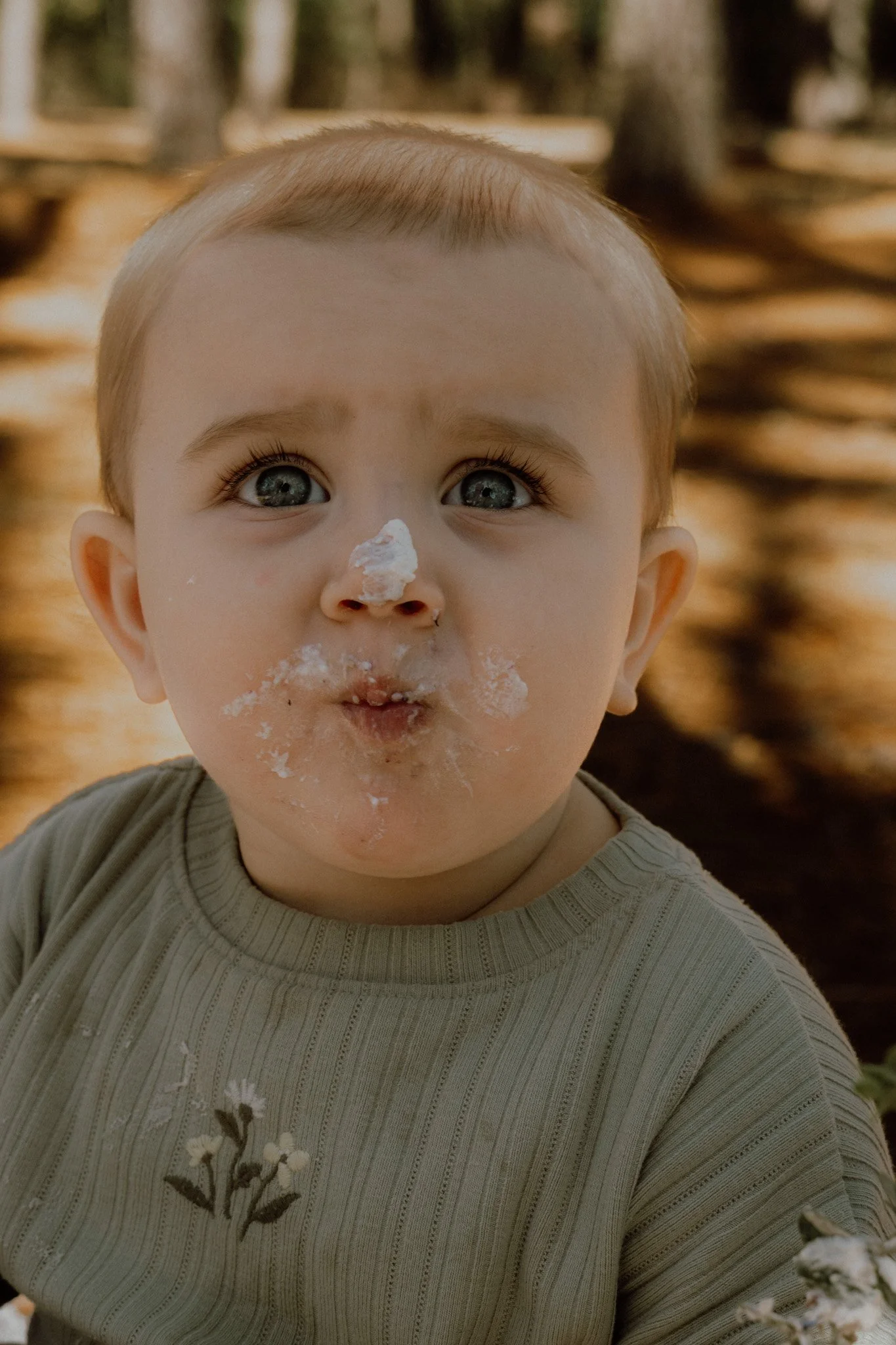 A young child with blue eyes and light brown hair, covered in white powder around the nose and mouth, wearing a beige shirt with a floral pattern, with a blurry natural background.