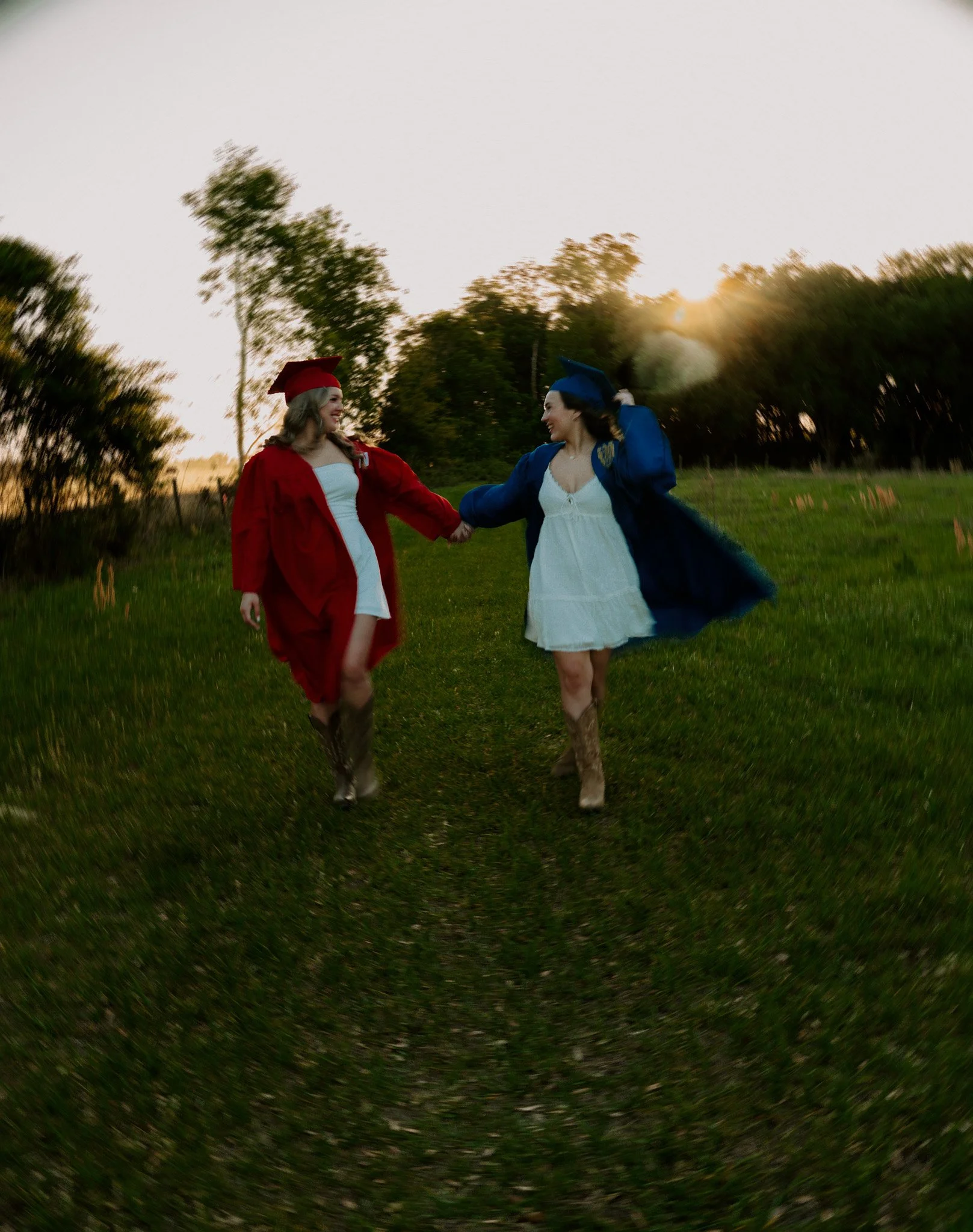Two women wearing graduation caps and gowns holding hands and walking through a grassy field during sunset.