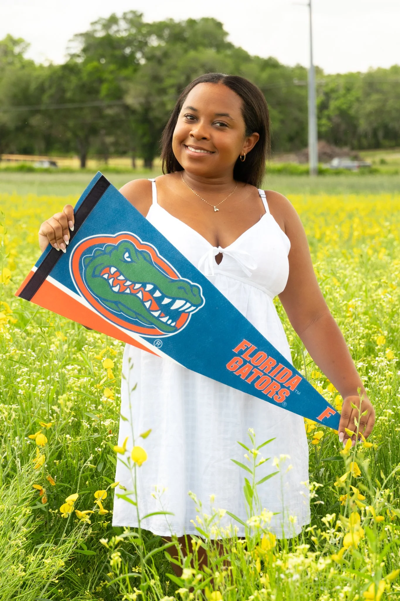 A woman standing in a field of yellow flowers, holding a Florida Gators banner, with trees and power lines in the background.
