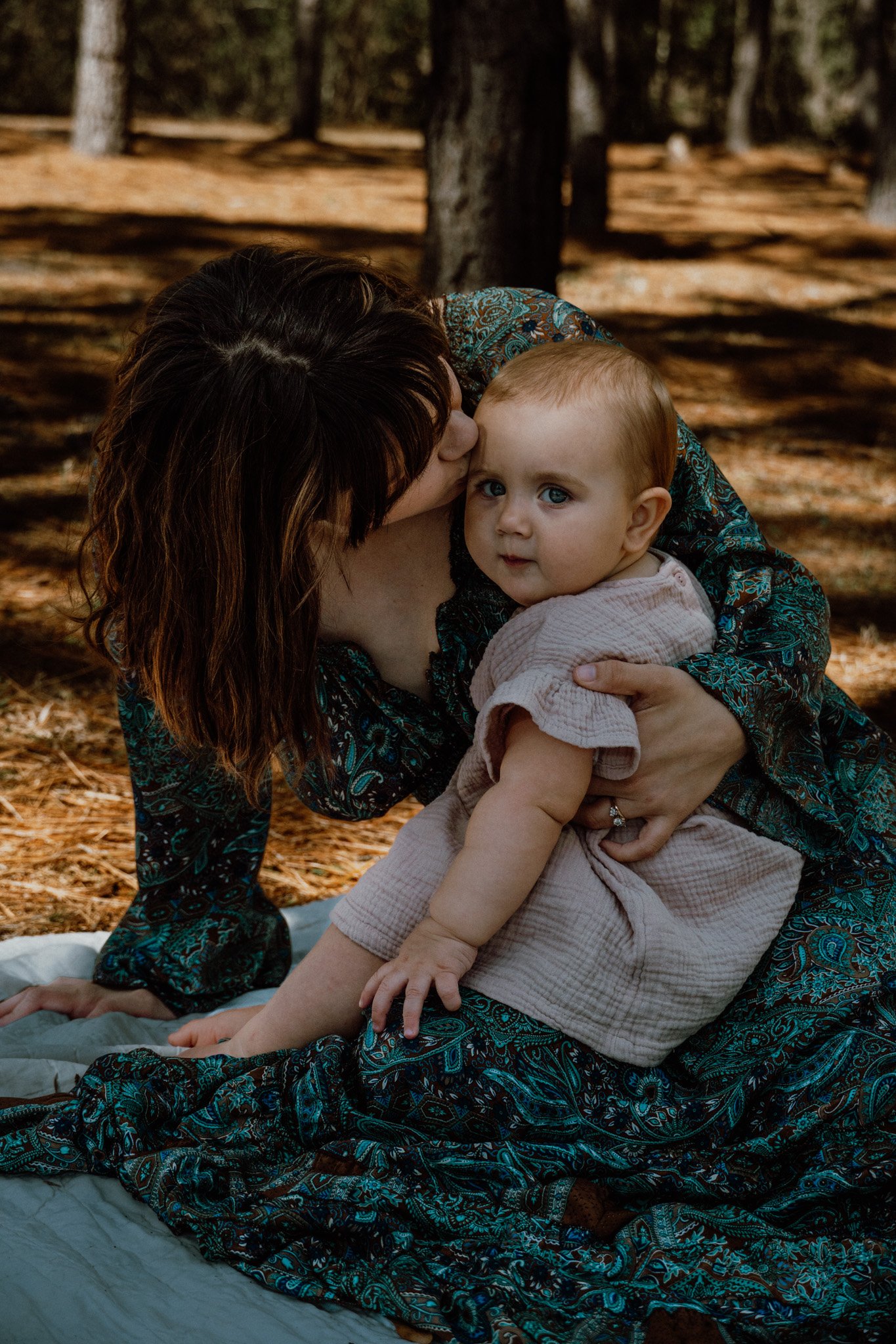 A woman gently kisses a young child's forehead while holding him in her arms outdoors in a wooded area with trees and a blanket on the ground.
