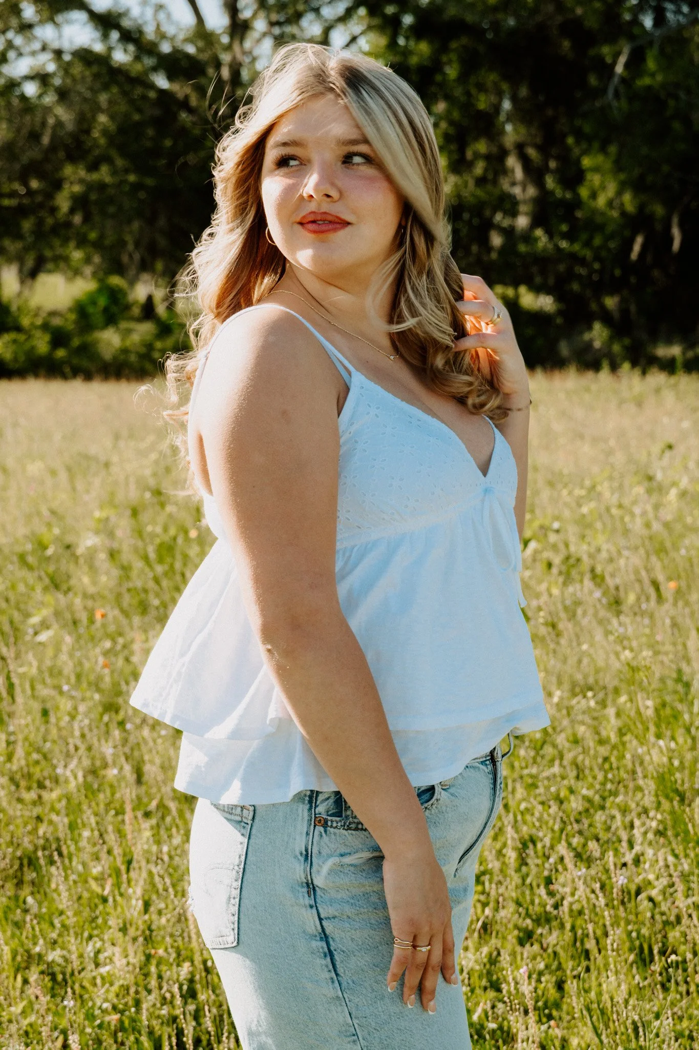 A young woman with long blonde hair wearing a white sleeveless top and light blue jeans, standing outdoors in a grassy field with trees in the background.
