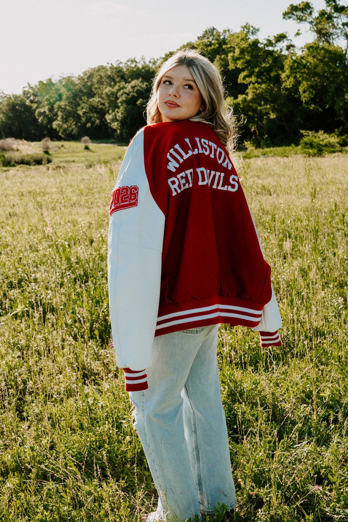 A young woman with blonde hair standing in a grassy field with trees in the background, wearing a red and white varsity jacket with team name and number patches, light-colored jeans, and striped socks.
