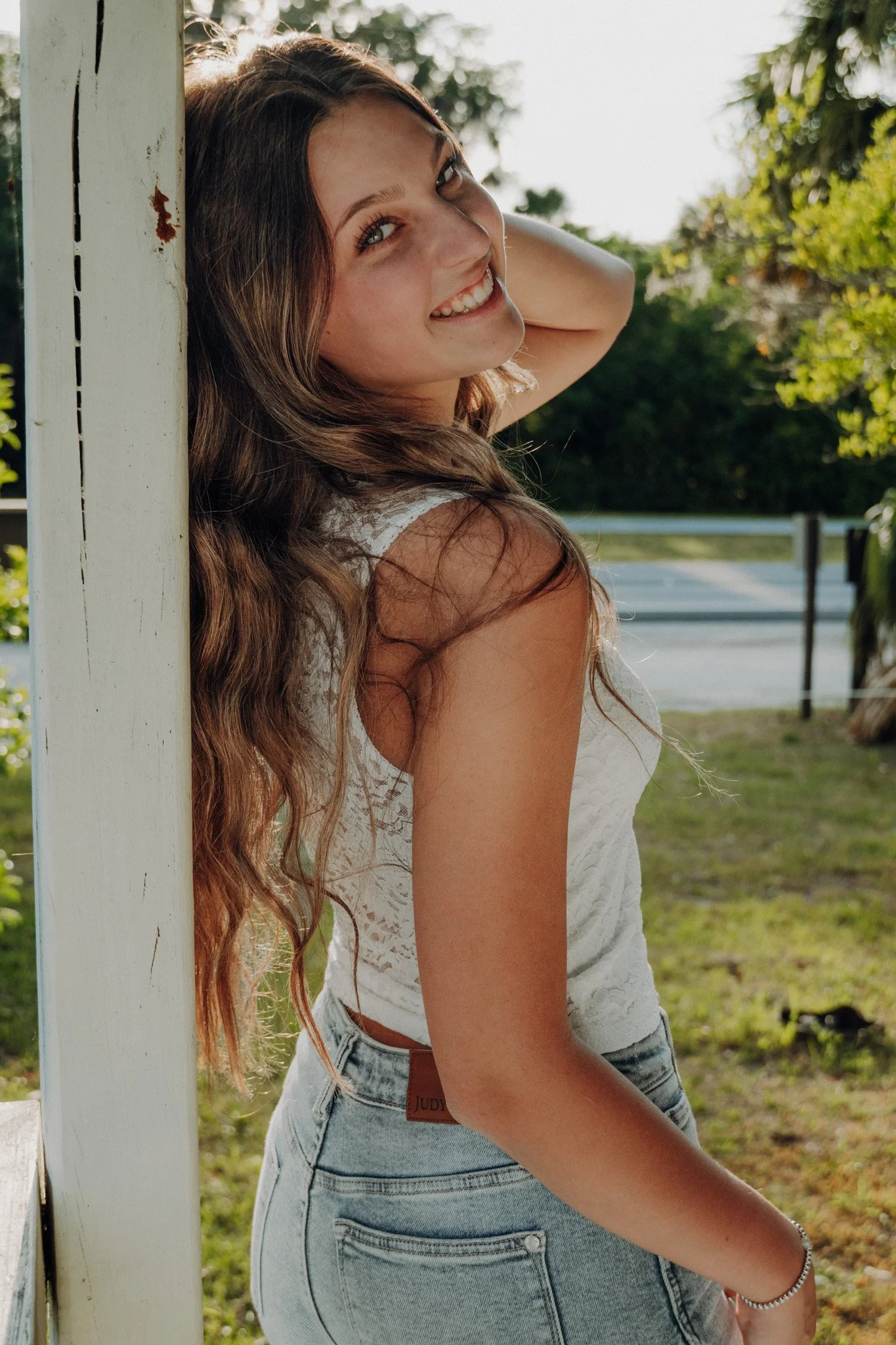 A young woman with long curly brown hair, smiling, leaning her head against a white wooden post outdoors. She is wearing a white lace top and high-waisted jeans, with greenery and trees in the background.