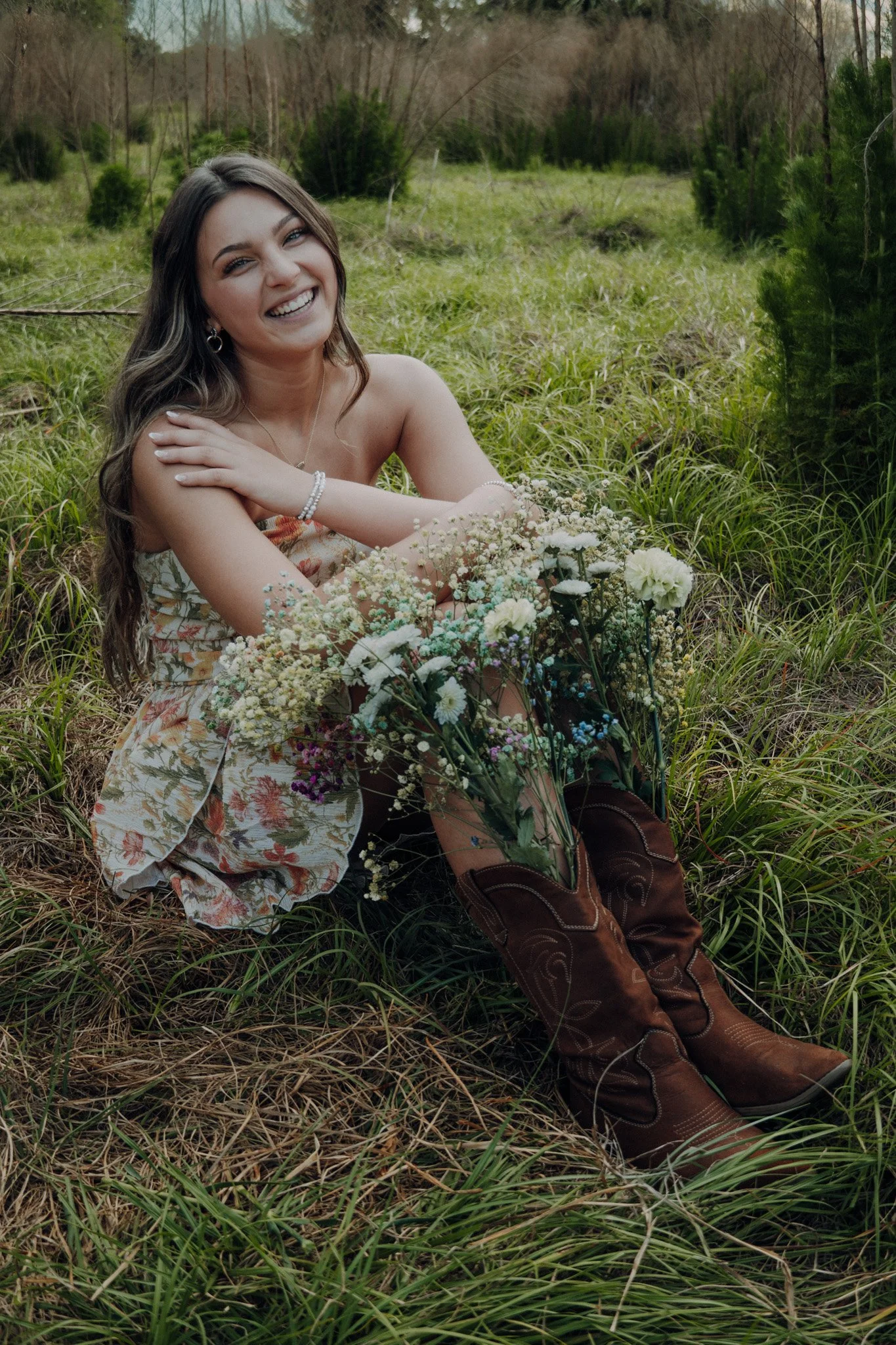 A smiling woman sitting in the grass with a bouquet of flowers, wearing a floral dress and cowboy boots, in an outdoor setting with greenery and trees.