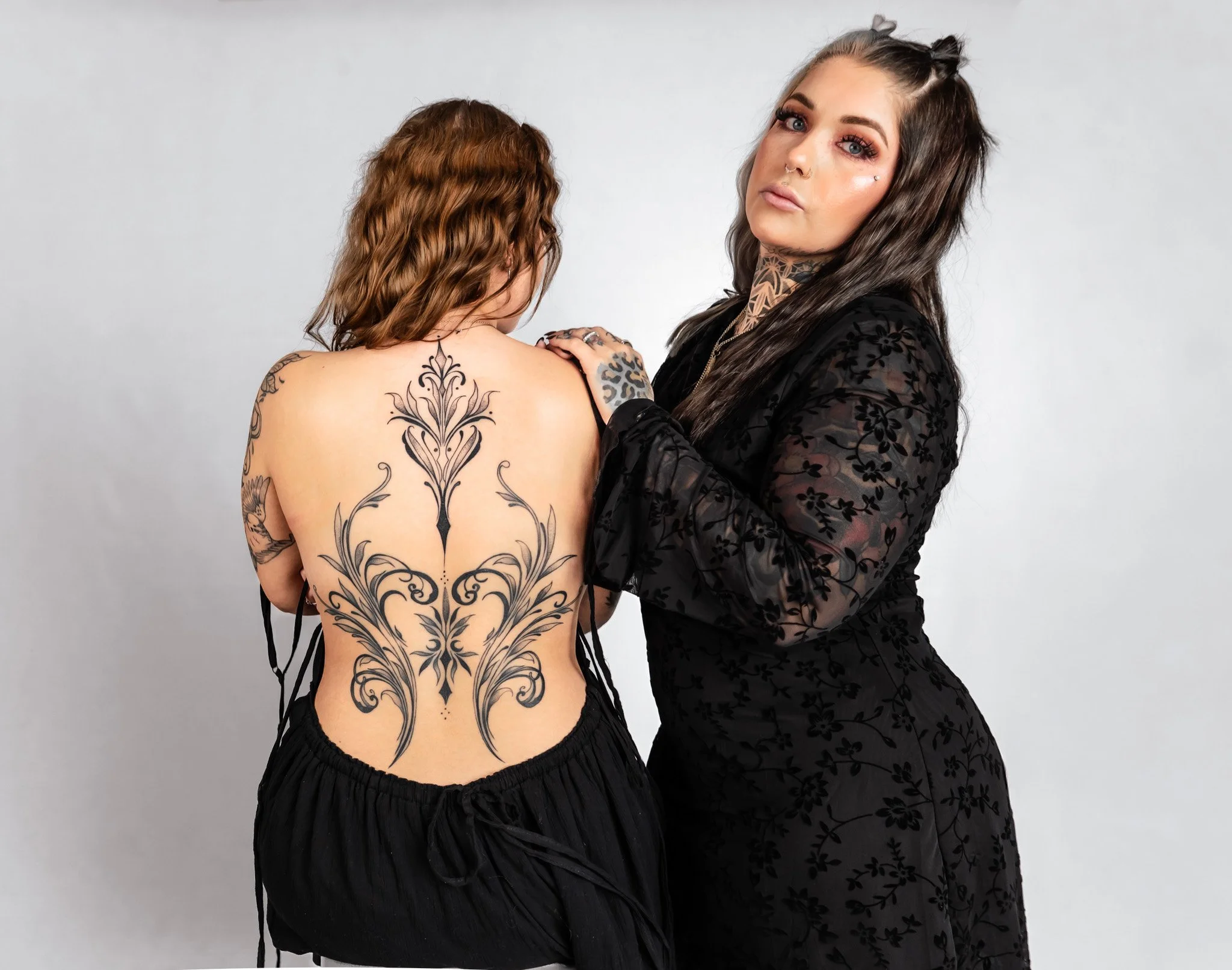 A woman with long, dark hair and tattoos on her neck and arms, wearing a black dress with floral patterns, stands next to another woman with shoulder-length brown hair, who has an intricate black tattoo on her back and is wearing black shorts, agains