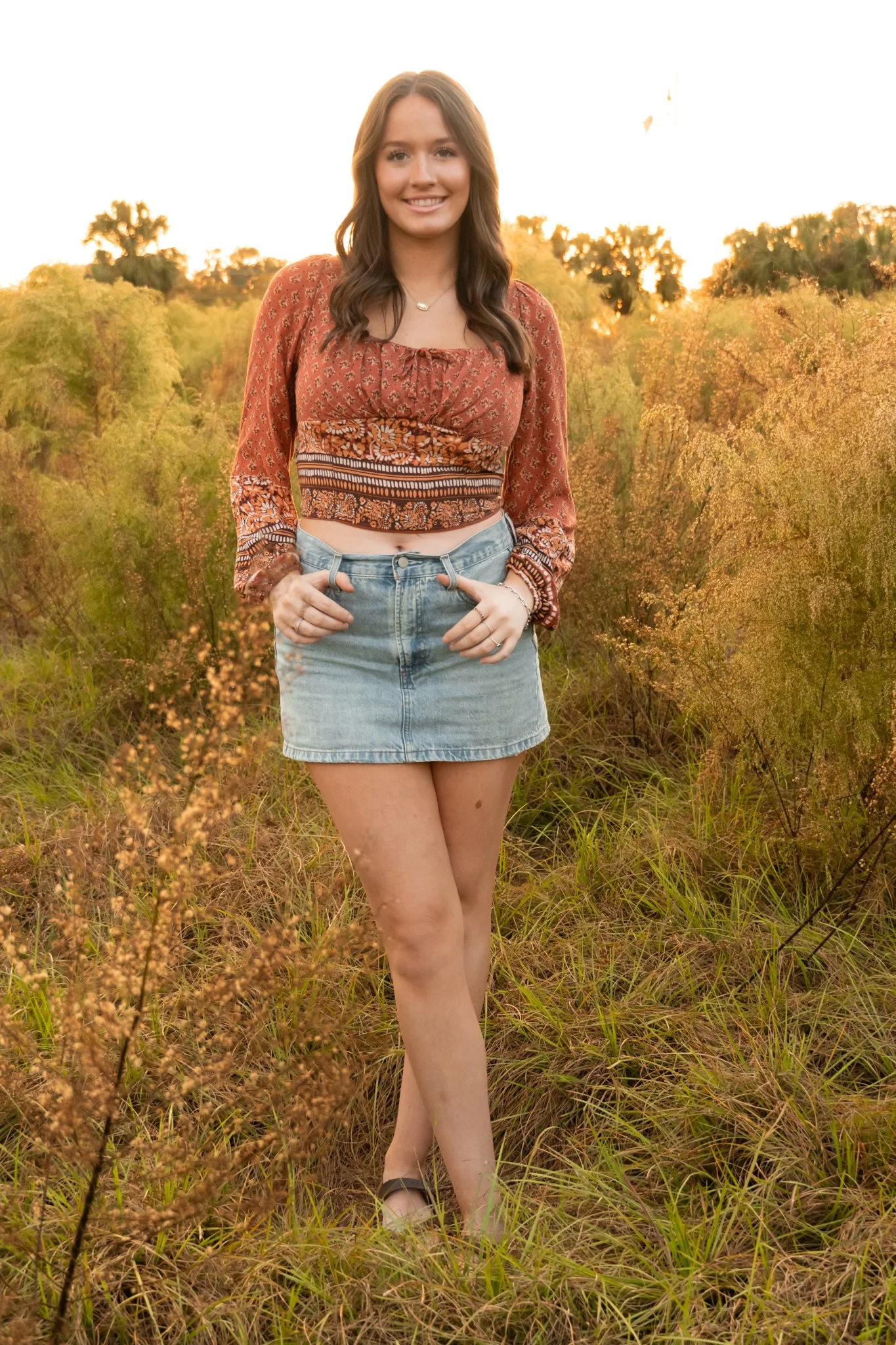 A young woman standing in a grassy field at sunset, smiling at the camera, wearing a patterned crop top, denim skirt, and sandals.
