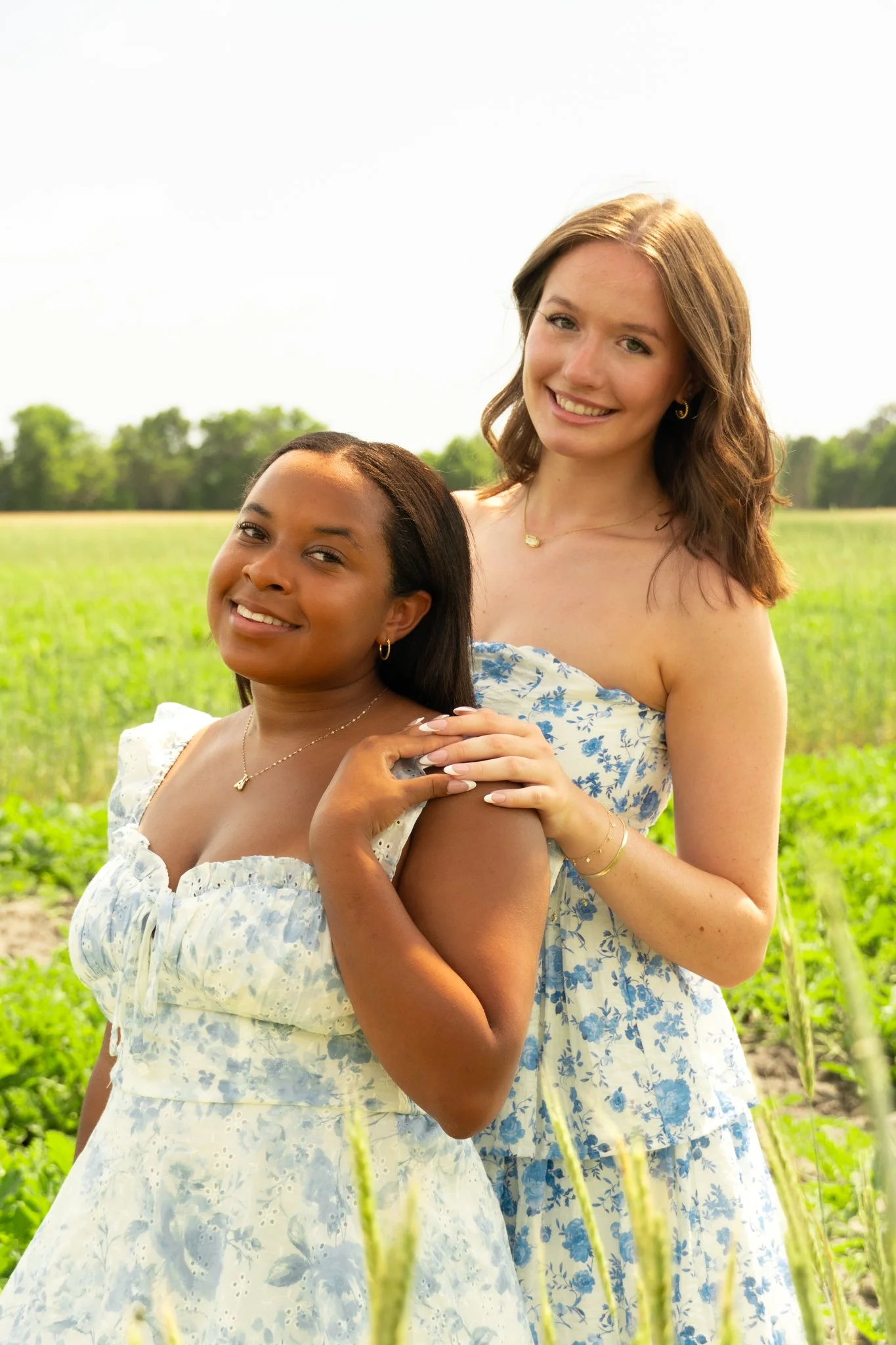 Two women standing close together outdoors in a green field, smiling at the camera, wearing light blue and white floral dresses, with trees in the background.